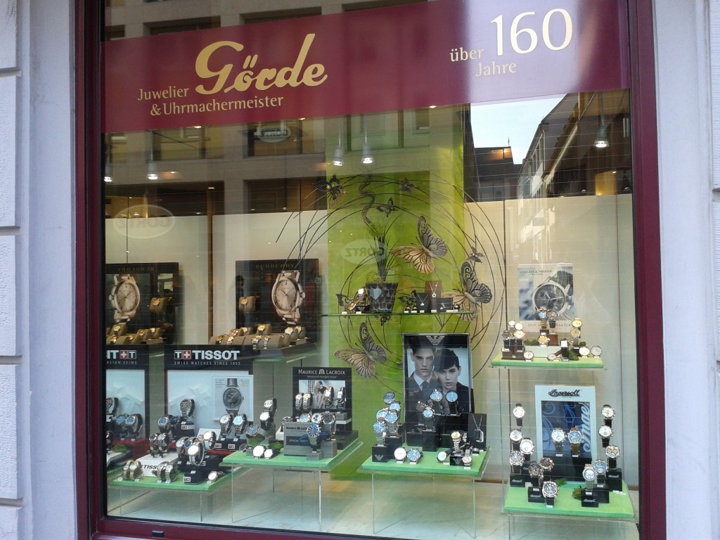 Schaufenster eines Juweliers mit Uhren und Schmuck, grüne Wandgestaltung mit Schmetterlingen, Marken wie Tissot und Maurice Lacroix sichtbar.