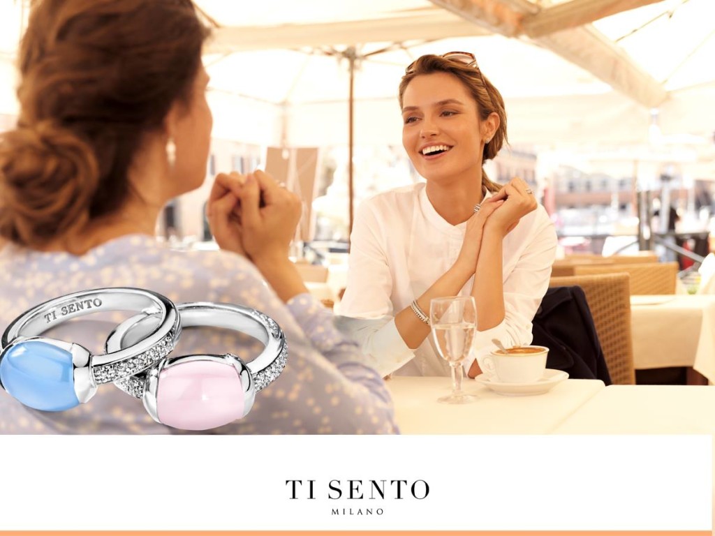 Zwei Frauen sitzen in einem Café. Eine lächelt und trägt ein weißes Oberteil, während die andere mit einem blauen Oberteil zuhört. Im Vordergrund sind zwei Ringe von TI SENTO zu sehen, einer blau und einer pink.
