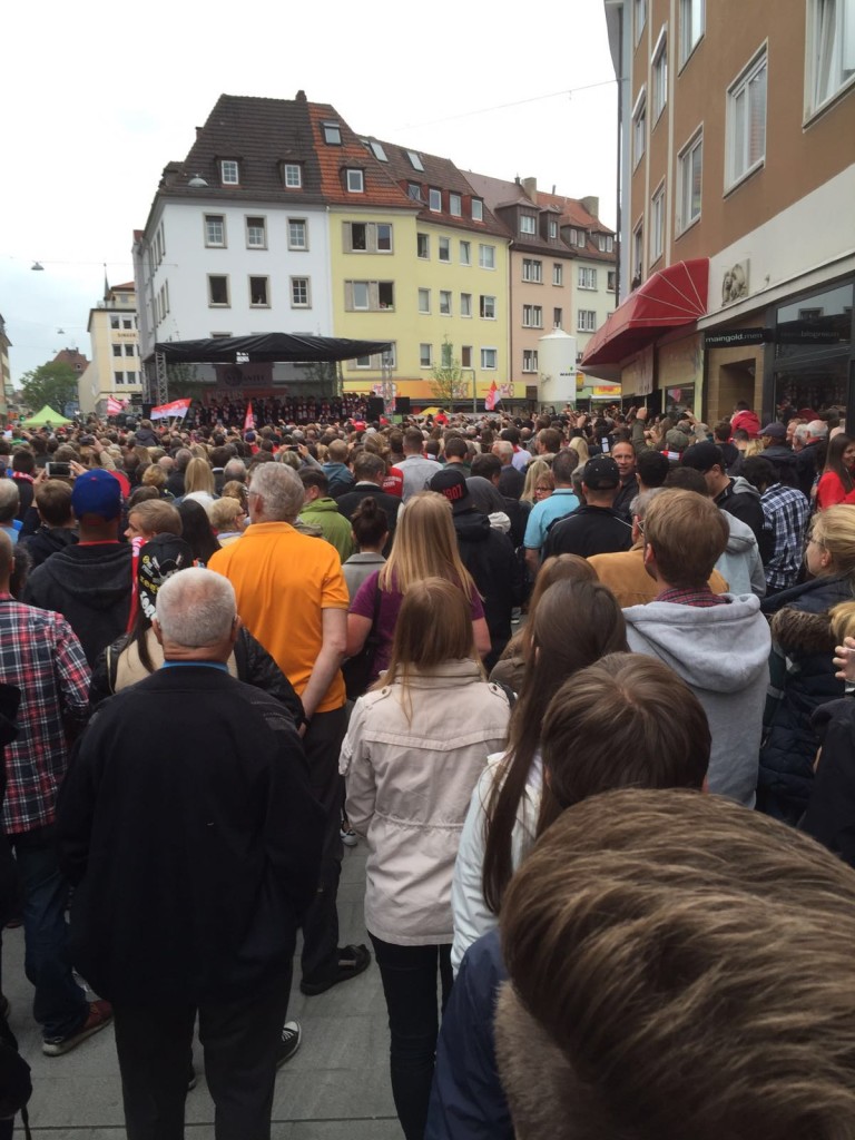 Eine große Menschenmenge versammelt sich im Stadtzentrum vor einer Bühne, umgeben von Gebäuden und Geschäften, bei einer Veranstaltung.