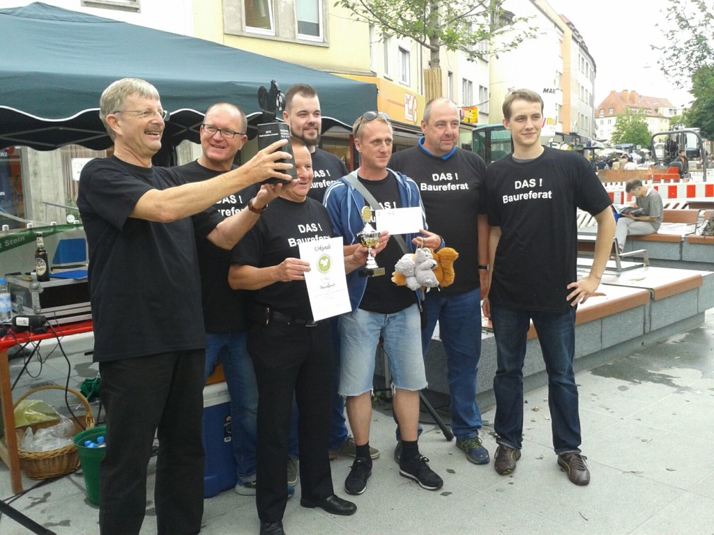 Sechs Männer in schwarzen T-Shirts mit dem Aufdruck "DAS ! Baureferat" posieren lächelnd für ein Gruppenfoto, während einer von ihnen ein Handy benutzt. Einige halten Pokale und Urkunden in der Hand, die Szene ist festlich und entspannt.