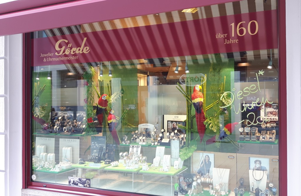 Schaufenster des Juweliers Göde mit Schmuck und Uhren in einer ansprechenden Ausstellung, dekoriert mit grünen Pflanzen und einem bunten Papageien.