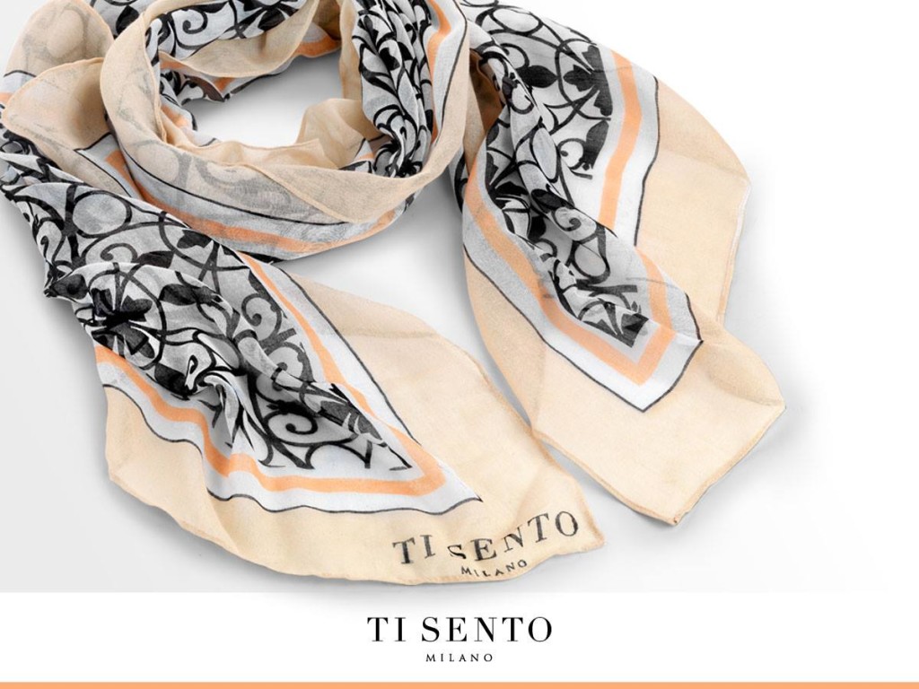 Ein leichter, beigefarbener Schal mit schwarzem, geschwungenem Muster und orangefarbener Umrandung. Am Ende ist das Logo "TI SENTO Milano" sichtbar.