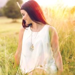 Eine Frau mit langen, dunklen Haaren trägt ein helles, fließendes Kleid und sitzt in hohem Gras, umgeben von sanften Sonnenstrahlen.