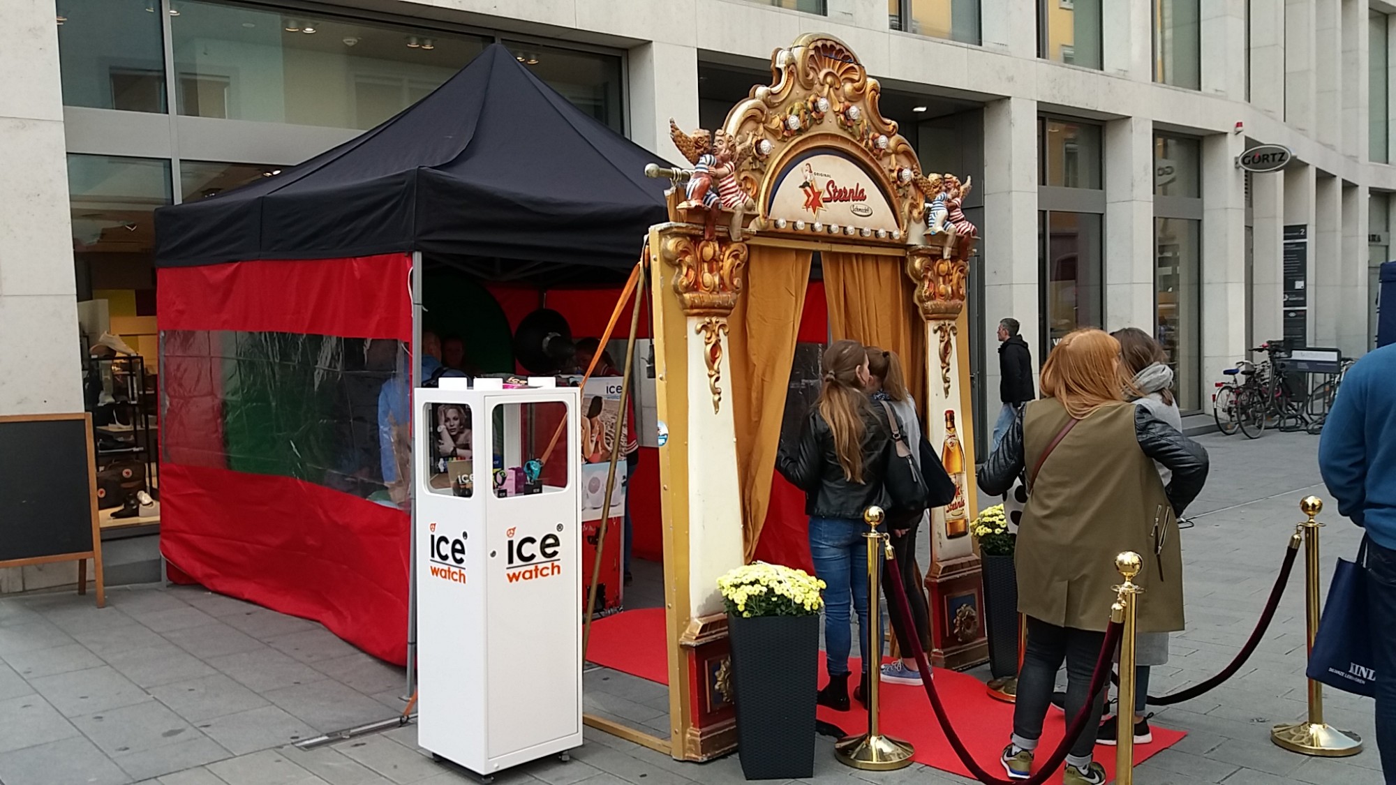 Ein festlicher Stand mit rotem Vorhang, einer goldenen Bühne und Seilen, vor denen Menschen auf Einlass warten. Nebenan steht ein Kiosk.