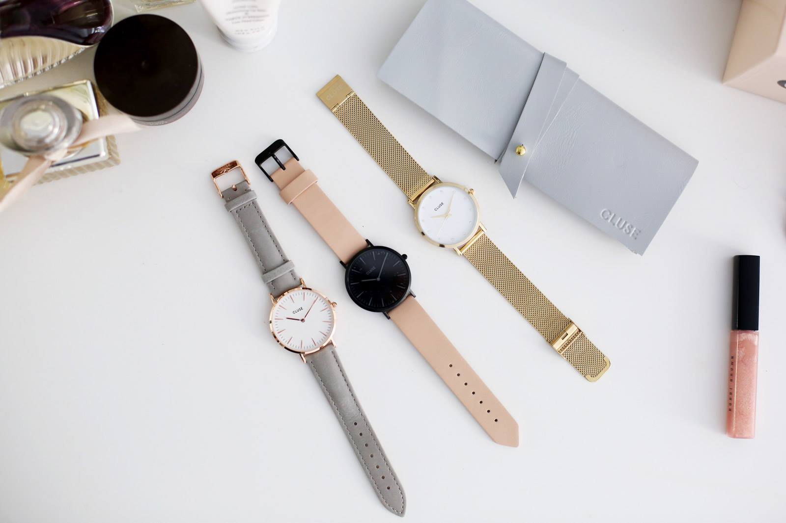 Drei stilvolle Armbanduhren in verschiedenen Designs liegen auf einem weißen Tisch, begleitet von Kosmetikprodukten und einem grauen Uhrenetui.