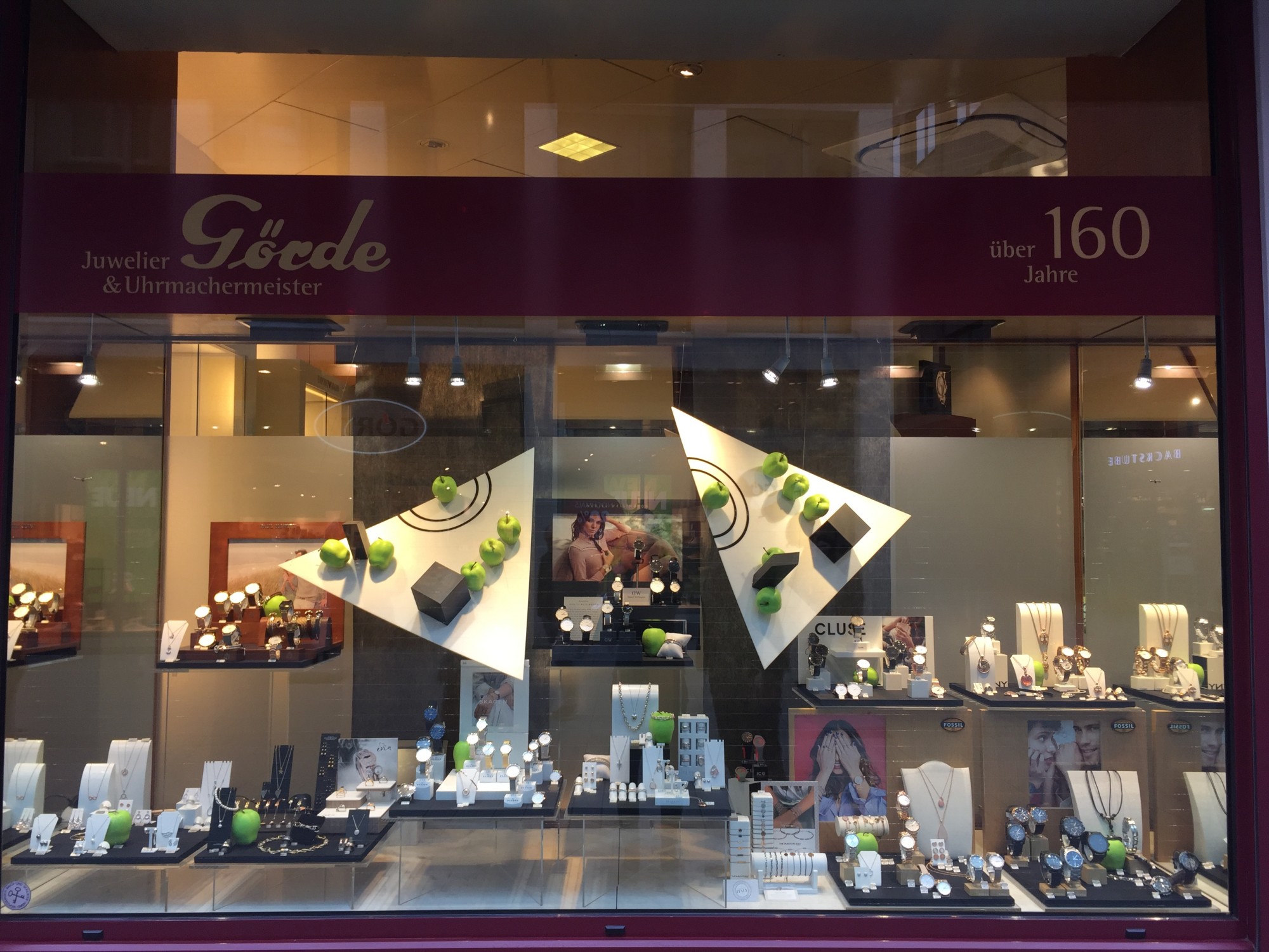 Schaufenster eines Juweliers mit modischen Schmuckstücken und Uhren, präsentiert auf eleganten Ständern, umrahmt von grünen Äpfeln.