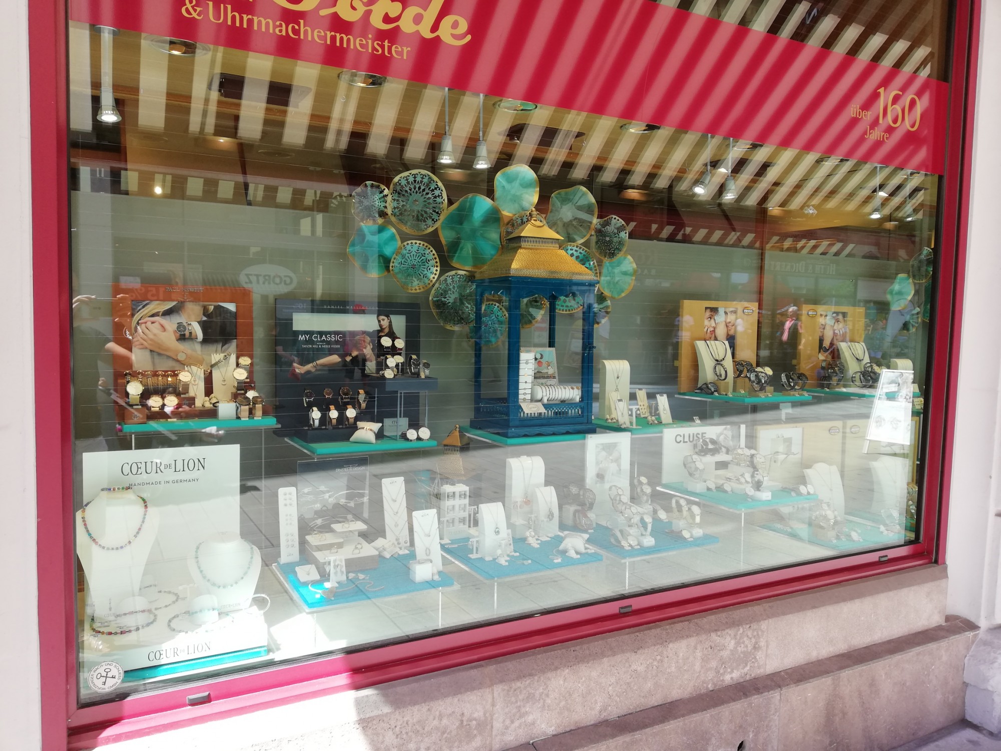 In einem Schaufenster sind Schmuck und Uhren ausgestellt. Zu sehen sind Armbänder, Halsketten und verschiedene Uhren in einer attraktiven Präsentation.