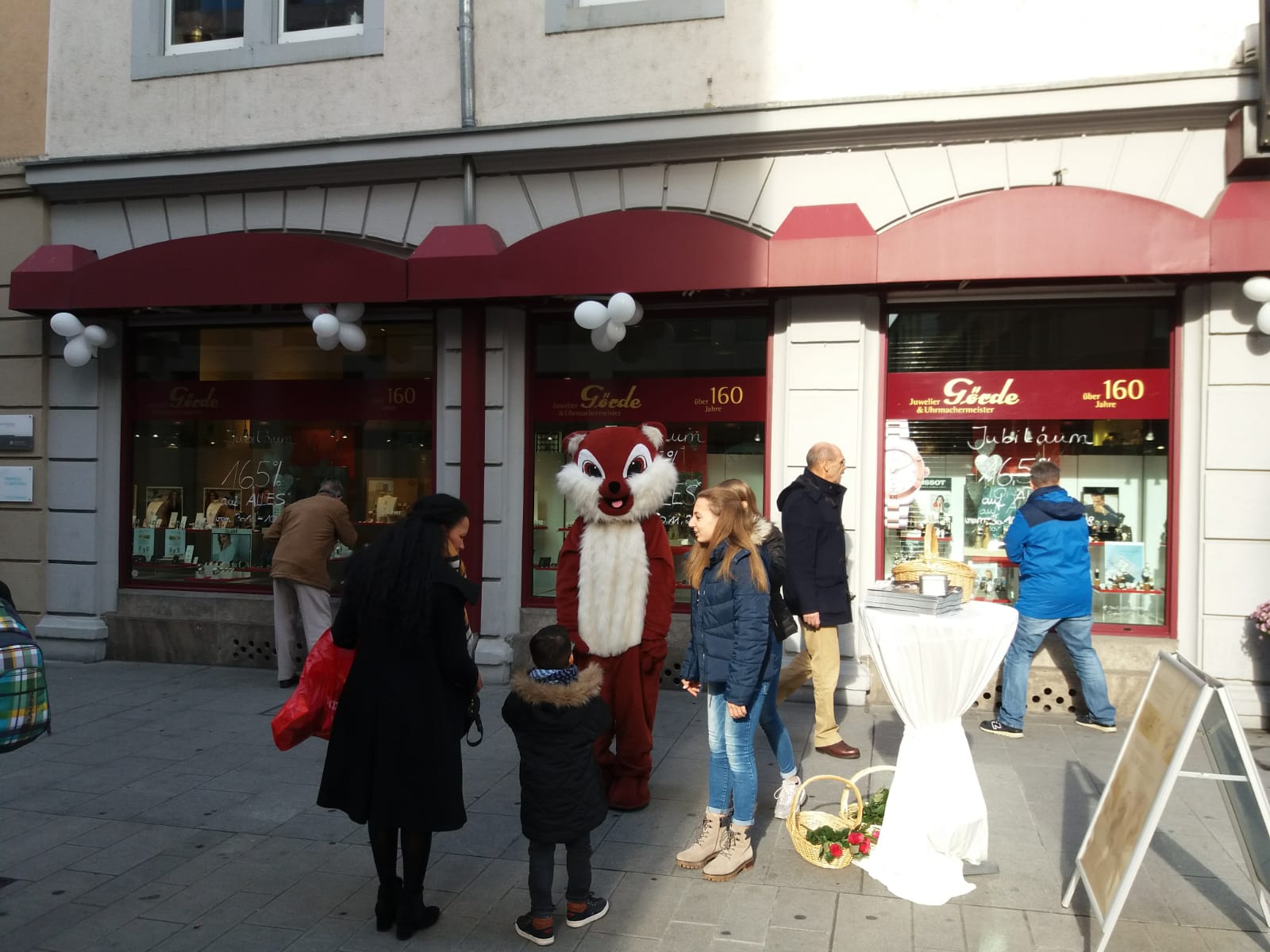 Auf einem Gehweg stehen mehrere Personen, während ein als Tier verkleideter Mensch in der Nähe eines Juweliergeschäfts lächelt. Im Hintergrund sind Schaufenster mit Dekoration zu sehen.