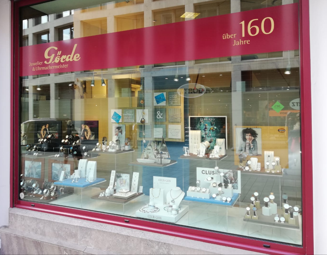 Das Schaufenster eines Juweliers zeigt eine Vielzahl von Schmuckstücken und Uhren. Im Hintergrund sind Werbefotos und Schriftzüge sichtbar.