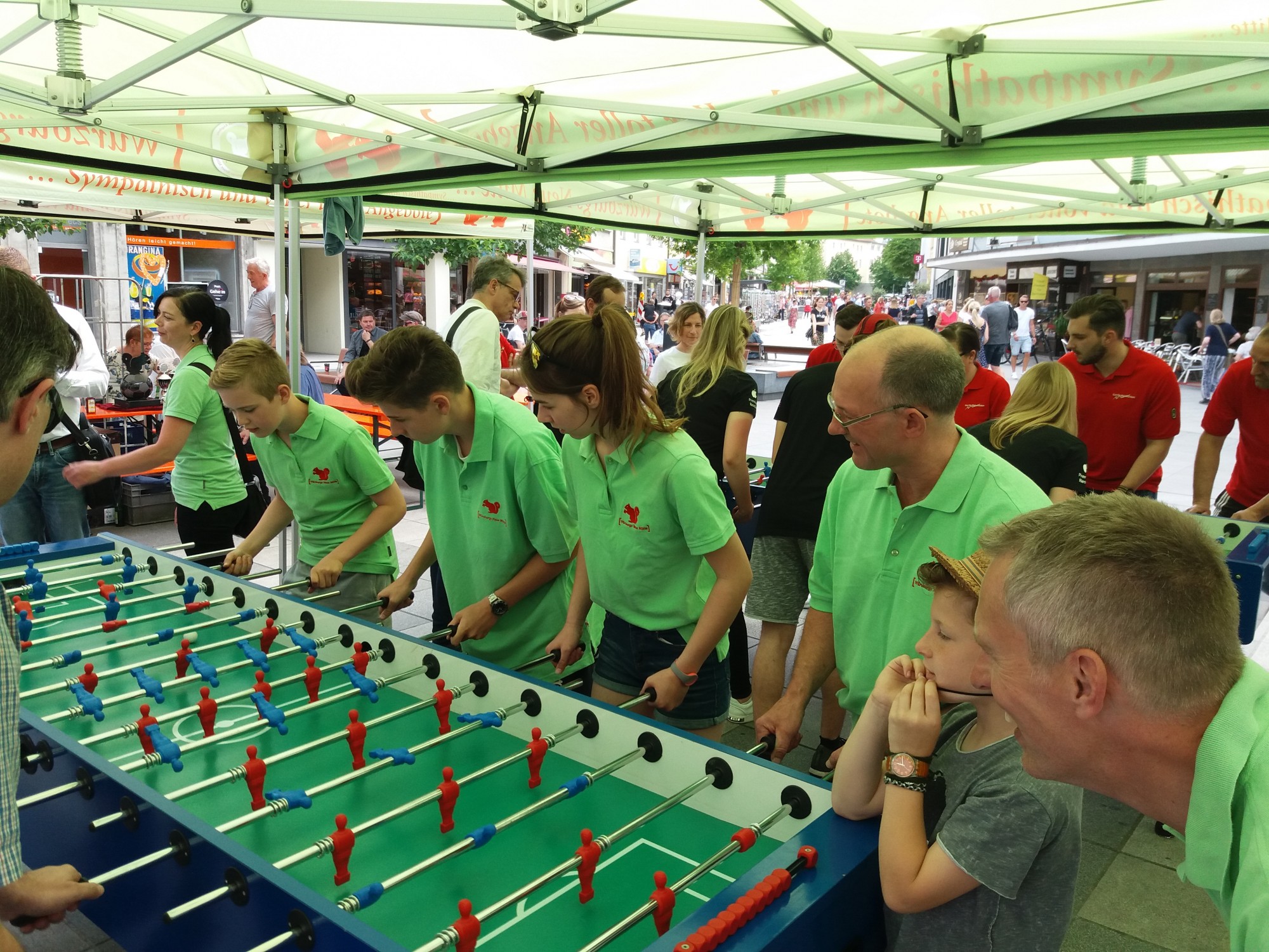 Eine Gruppe von Menschen in den Farben Grün und Rot spielt Tischfußball unter einem Zelt. Einige Zuschauer beobachten gespannt das Geschehen.