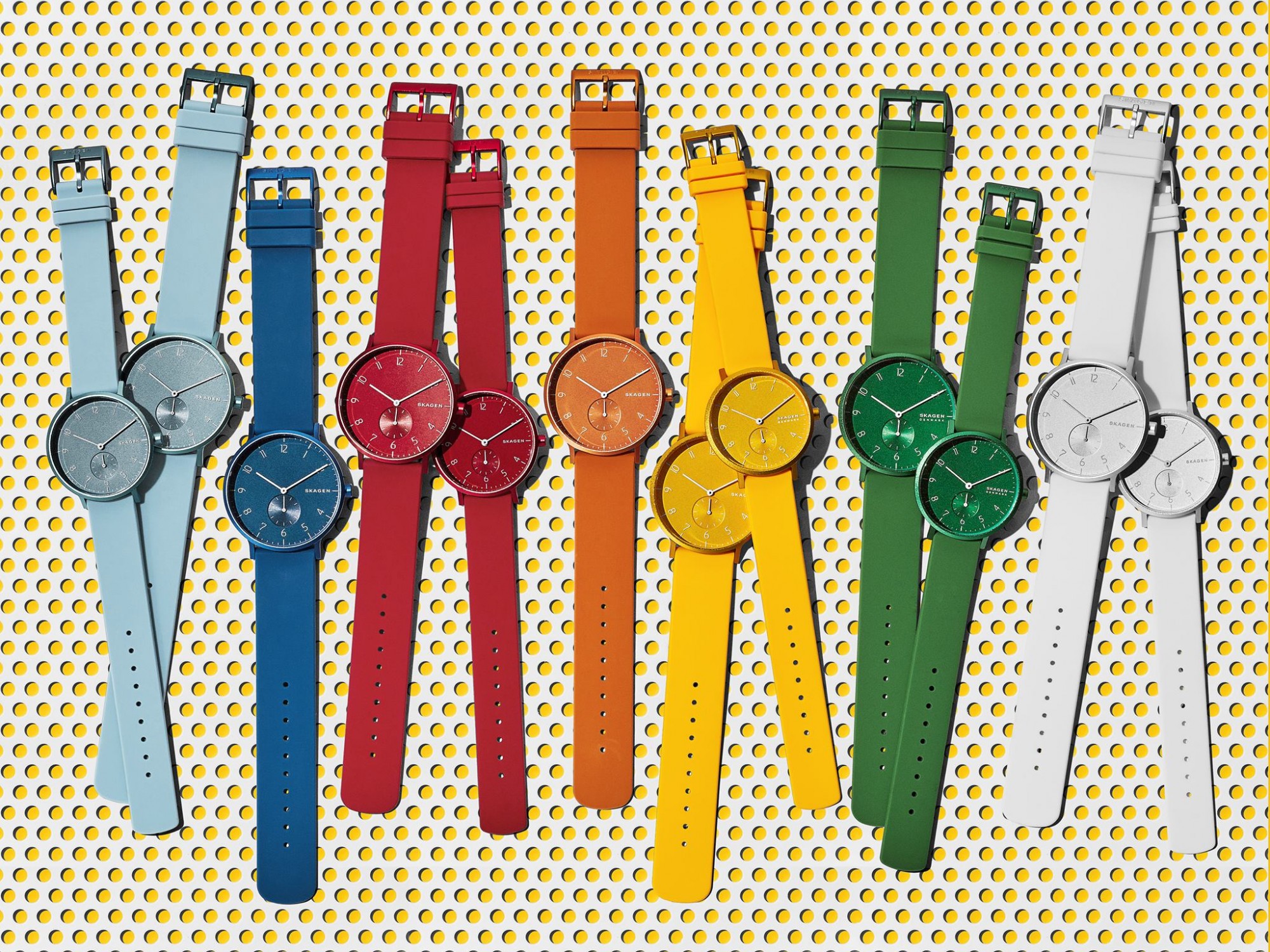 Eine Vielzahl von Uhren in verschiedenen Farben mit farblich abgestimmten Armbändern, angeordnet auf einer gelben, gepunkteten Unterlage.