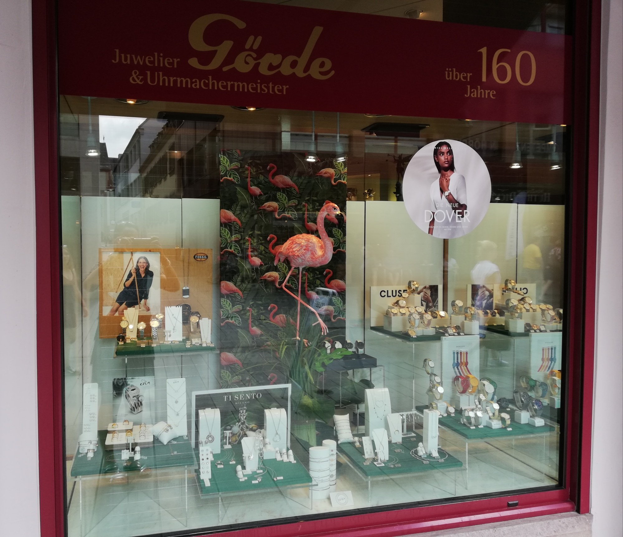Die Schaufensterfront eines Juweliers zeigt verschiedene Schmuckstücke und Uhren. Im Hintergrund ist ein farbenfrohes Flamingo-Motiv zu sehen.
