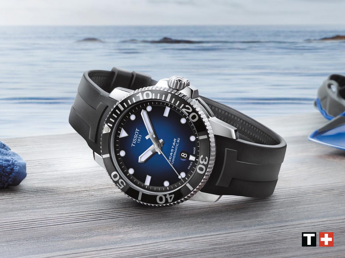 Eine elegante Tissot Taucheruhr mit schwarzem Silikonarmband liegt auf einer Holzoberfläche. Im Hintergrund ist ein Meer mit sanften Wellen zu sehen.