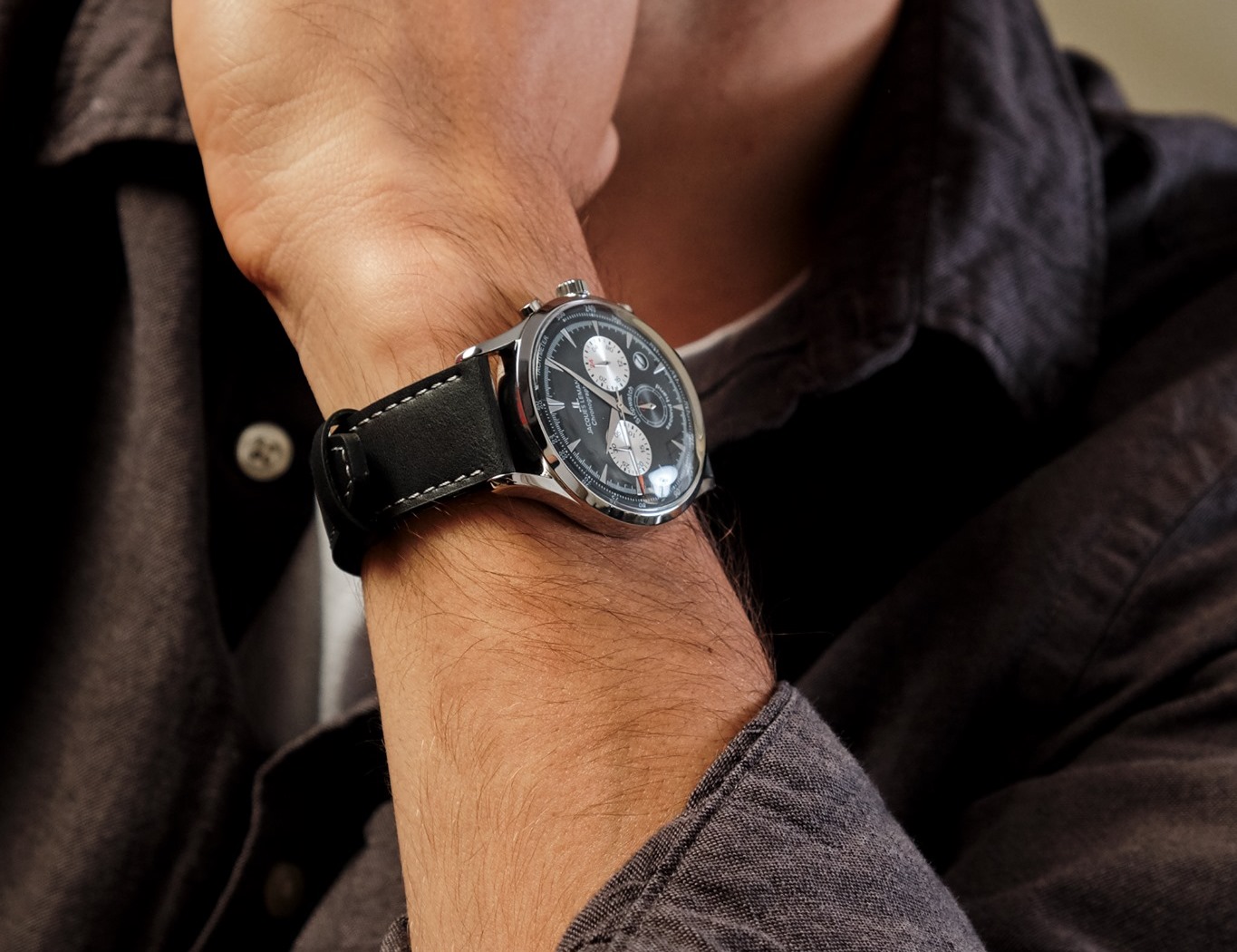 Eine nahaufgenommene Hand trägt eine elegante Armbanduhr mit schwarzem Lederarmband und dunklem Ziffernblatt, mit mehreren Ziffernblättern in einer stilvollen Umgebung.