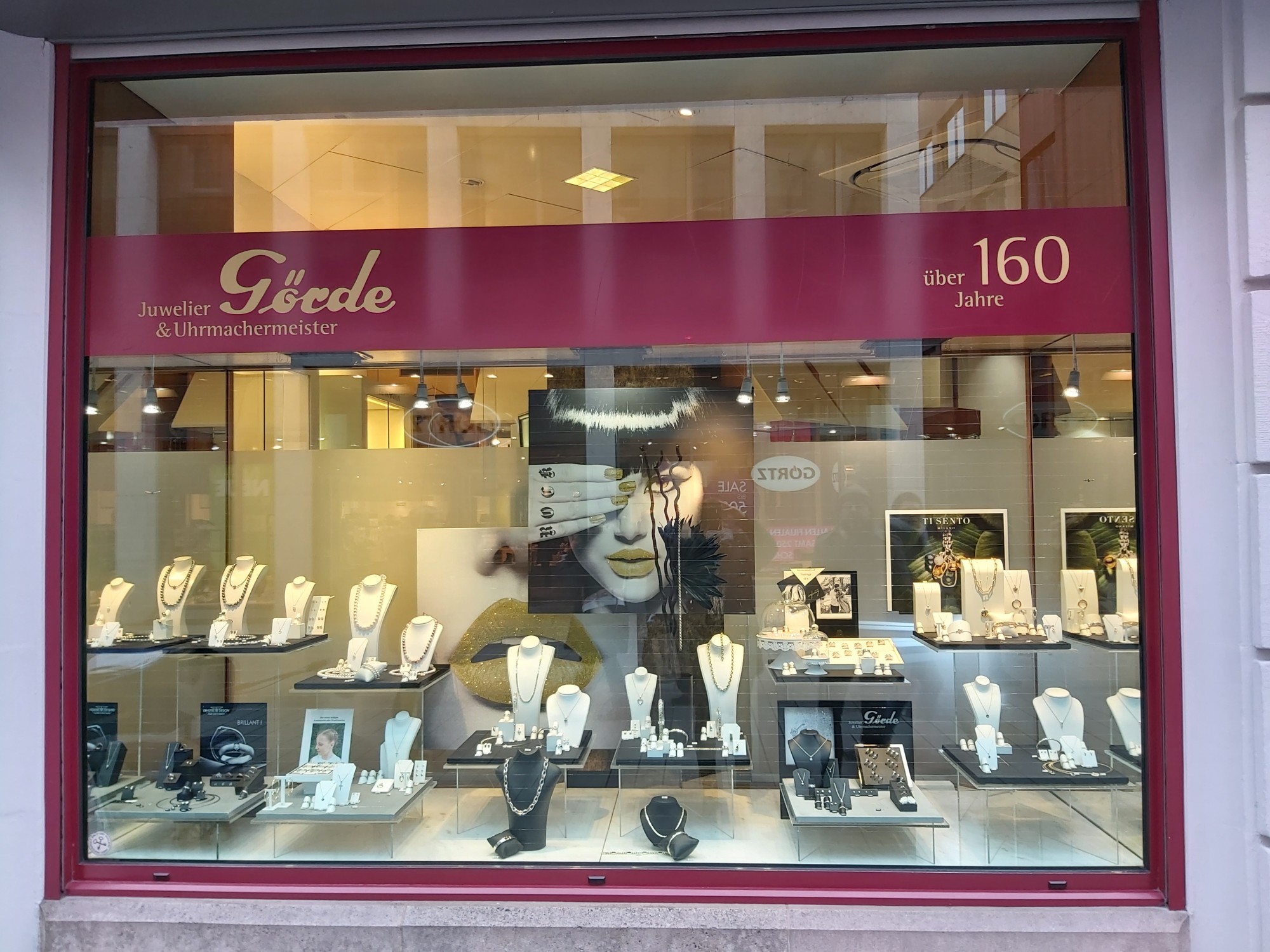 Das Bild zeigt die Schaufensterfront eines Juweliergeschäfts mit eleganten Schmuckexponaten, einer großen Werbung für den Laden und einer stilvollen Präsentation.