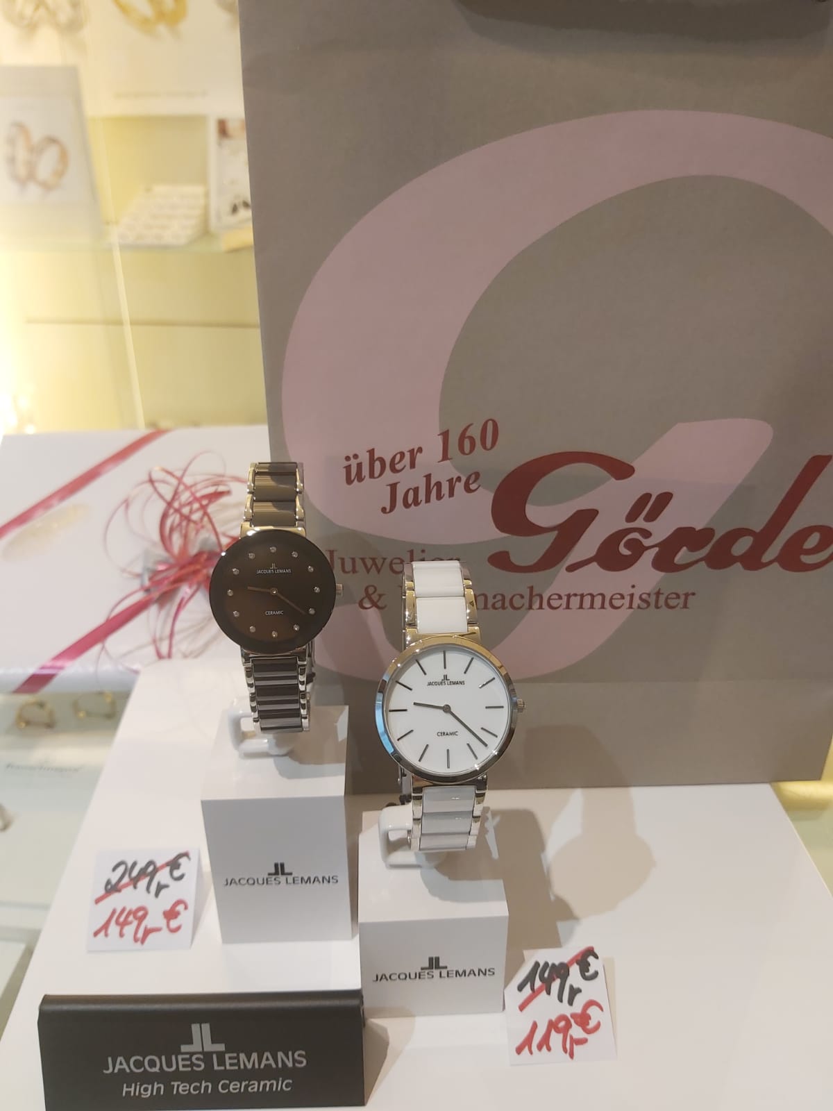 Zwei elegante Armbanduhren von Jacques Lemans stehen auf einem Tisch. Eine Uhr hat ein schwarzes Ziffernblatt, die andere ein weißes. Im Hintergrund ist eine Tüte mit dem Logo eines Juweliers zu sehen.
