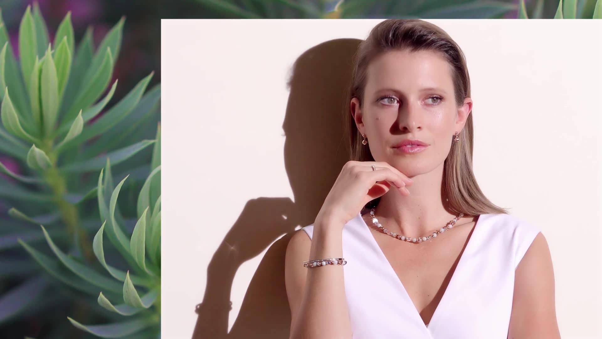 Eine Frau mit blonden Haaren in einem weißen Oberteil posiert elegant, während Schatten und sanfte Pflanzen im Hintergrund zu sehen sind.