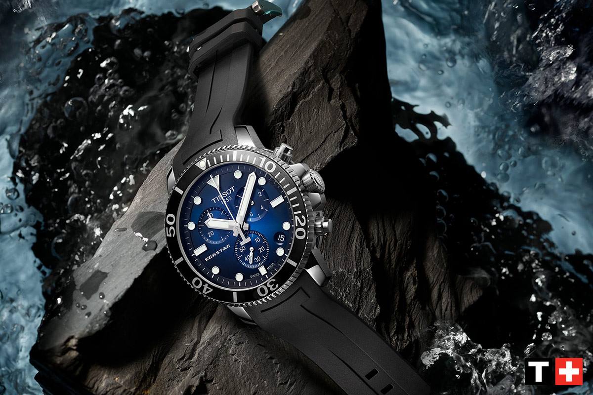Eine sportliche Armbanduhr von Tissot mit blauen Ziffern auf einem schwarzen Zifferblatt, platziert auf einem dunklen Stein, umgeben von spritzendem Wasser.