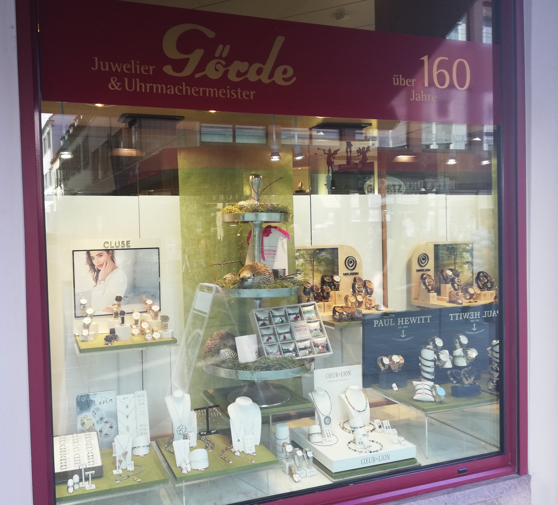 Schaufenster eines Juweliers mit Schmuck und Uhren. Die Präsentation zeigt verschiedene Marken, darunter Paul Hewitt und Cluse, in stilvollem Ambiente.