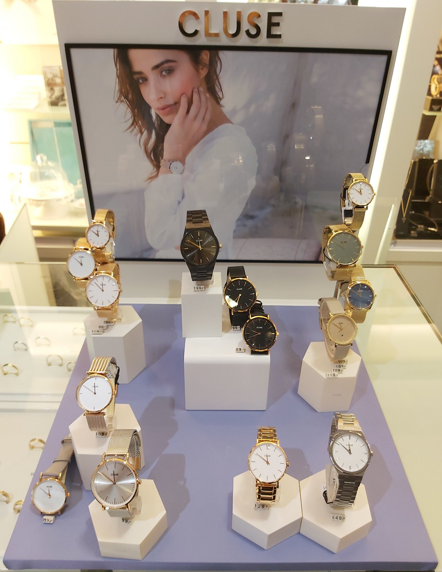 Eine stilvolle Präsentation verschiedener Cluse-Uhren auf einem Verkaufsdisplay, hinter dem ein Bild einer Frau mit einer Uhr zu sehen ist.