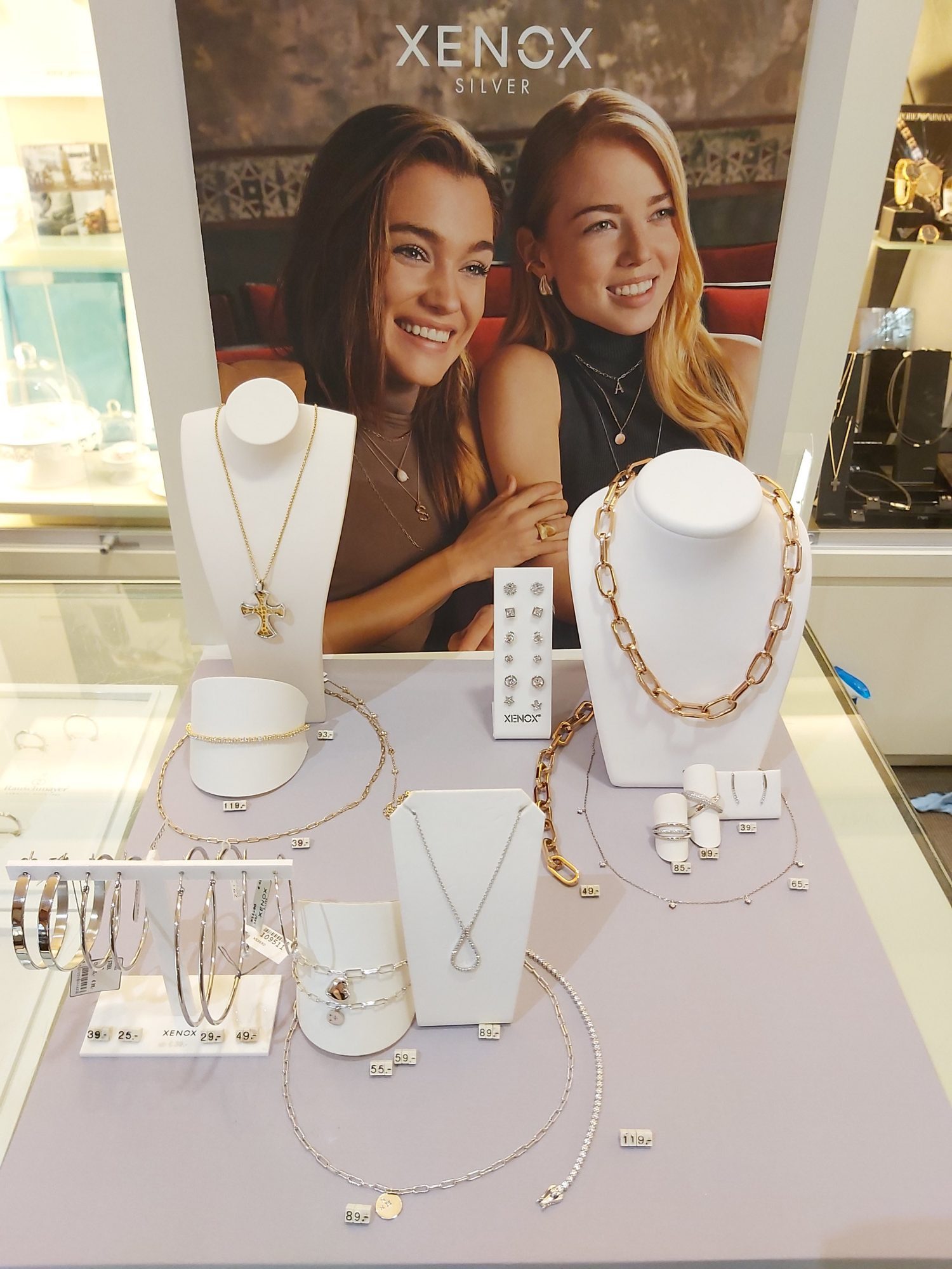 Das Bild zeigt eine Schmuckauslage mit verschiedenen Halsketten, Armbändern und Ohrringen. Im Hintergrund sind zwei lächelnde Frauen zu sehen, die modischen Schmuck tragen.