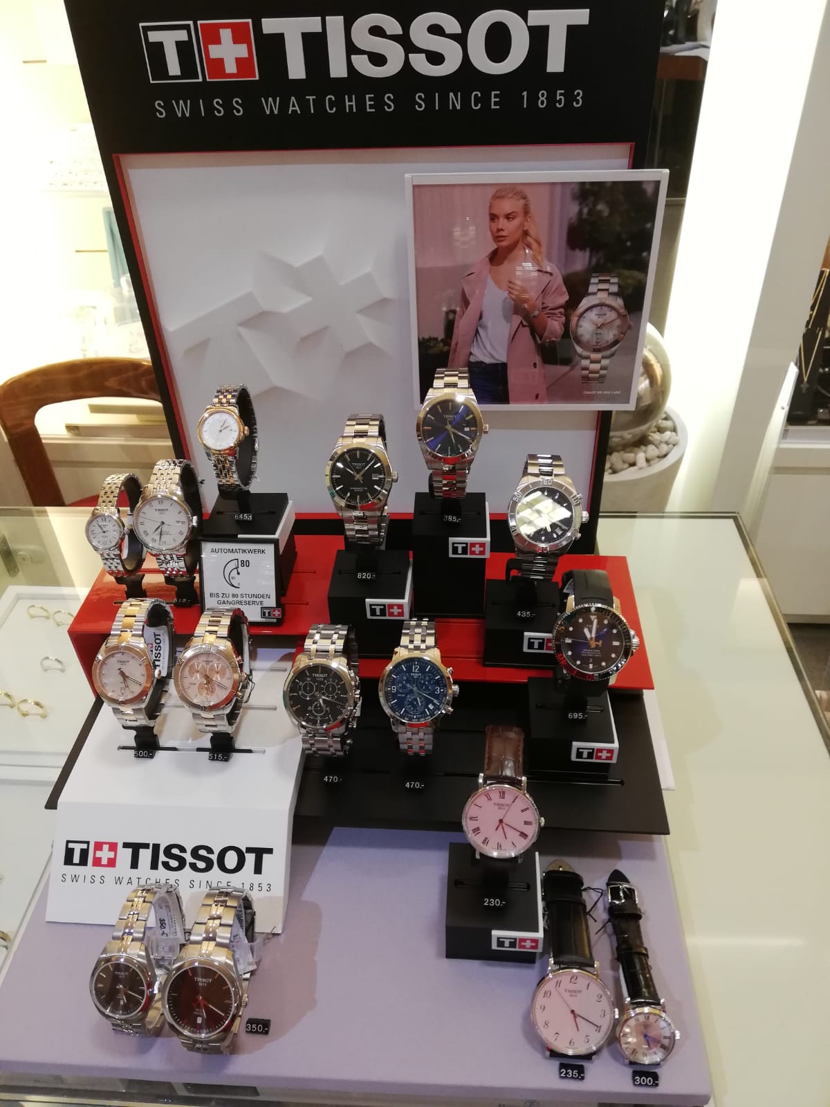 Eine Produktpräsentation von Tissot-Uhren in einem Geschäft. Mehrere Uhren in verschiedenen Designs und Farben sind in Vitrinen ausgestellt, begleitet von einer Werbefotografie.