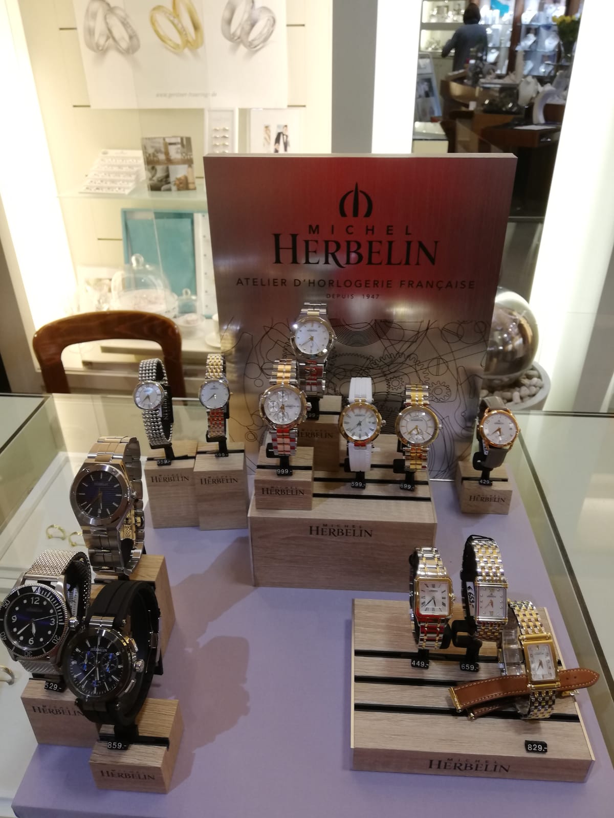 In einem eleganten Uhrenstand von Michel Herbelin sind verschiedene Armbanduhren auf Holzsockeln ausgestellt. Im Hintergrund sind weitere Schmuckstücke sichtbar.
