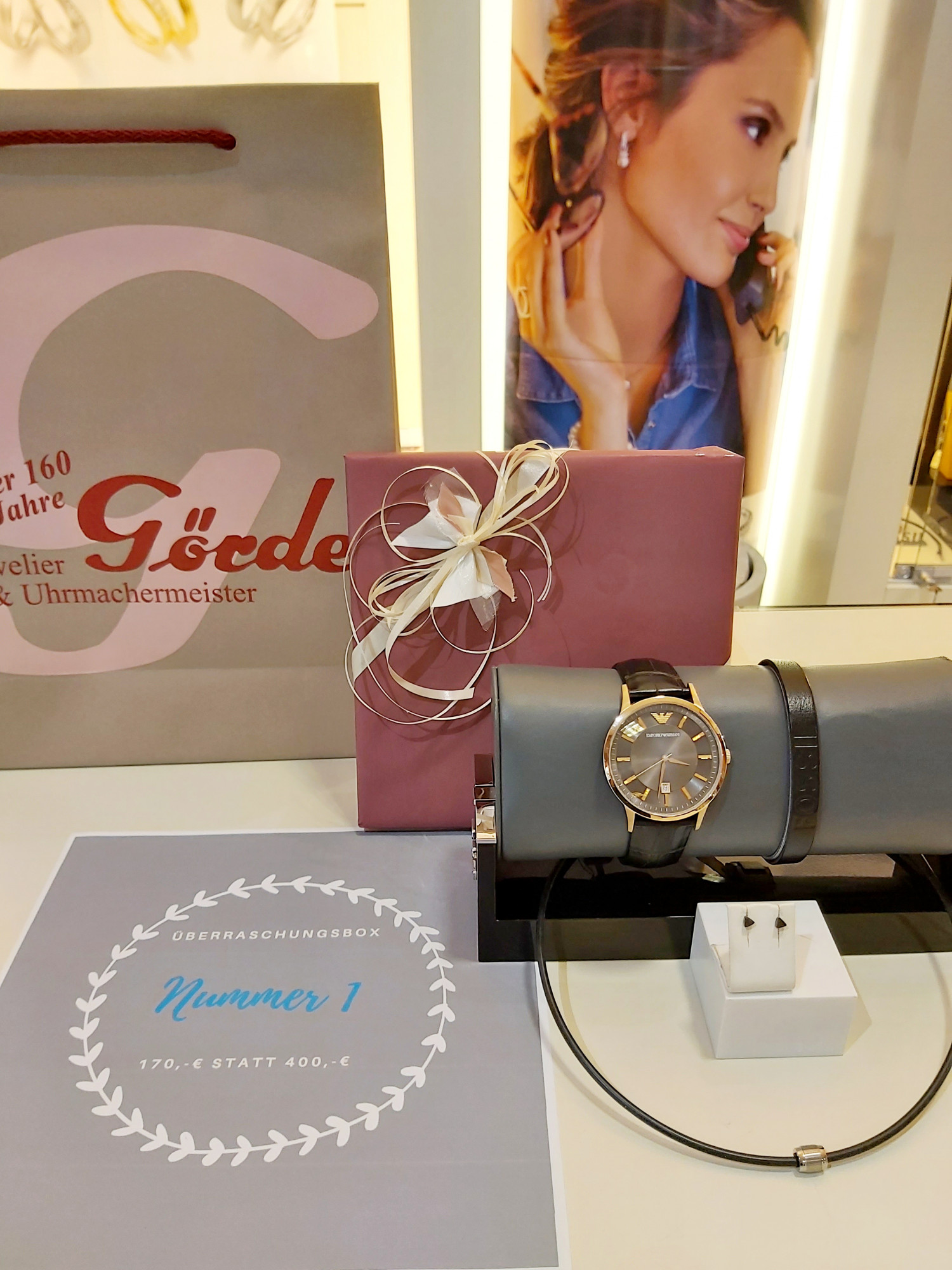 Eine elegante Armbanduhr und Schmuck in einer Überraschungsbox mit Preisangabe von 170 € statt 400 €, umgeben von Geschenkbändern und einer Einkaufstüte.