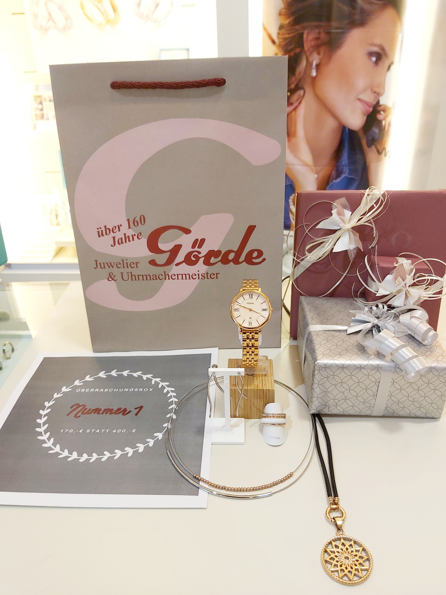 Ein dekorierter Tisch zeigt ein Geschenkpaket, einen Schmuckanhänger, eine Uhr und eine Broschüre mit einem Angebot für eine Überraschungsbox.