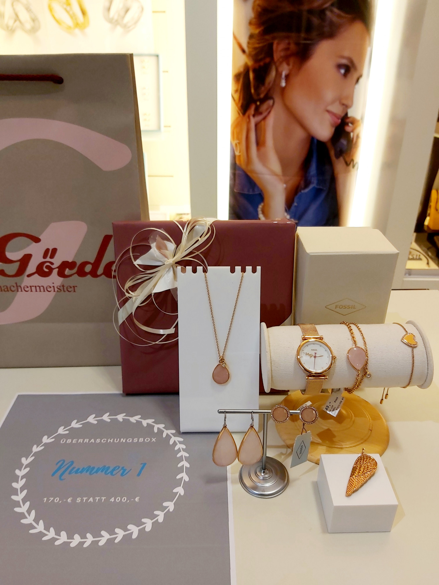 Im Bild sind verschiedene Schmuckstücke auf einem Tisch präsentiert, darunter eine Kette, Ohrringe und eine Uhr. Im Hintergrund ist eine Geschenkschachtel und ein Anzeigetafel mit Preisen zu sehen.