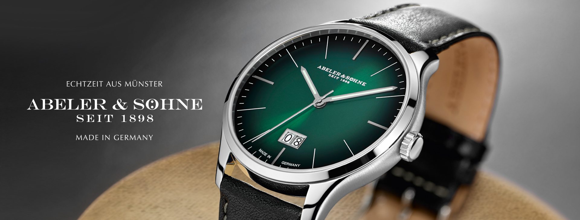 Eine elegante, grüne Armbanduhr von Abeler & Söhne mit einem schwarzen Lederarmband und einem Datumsauswahlfenster, platziert auf einer neutralen Oberfläche.