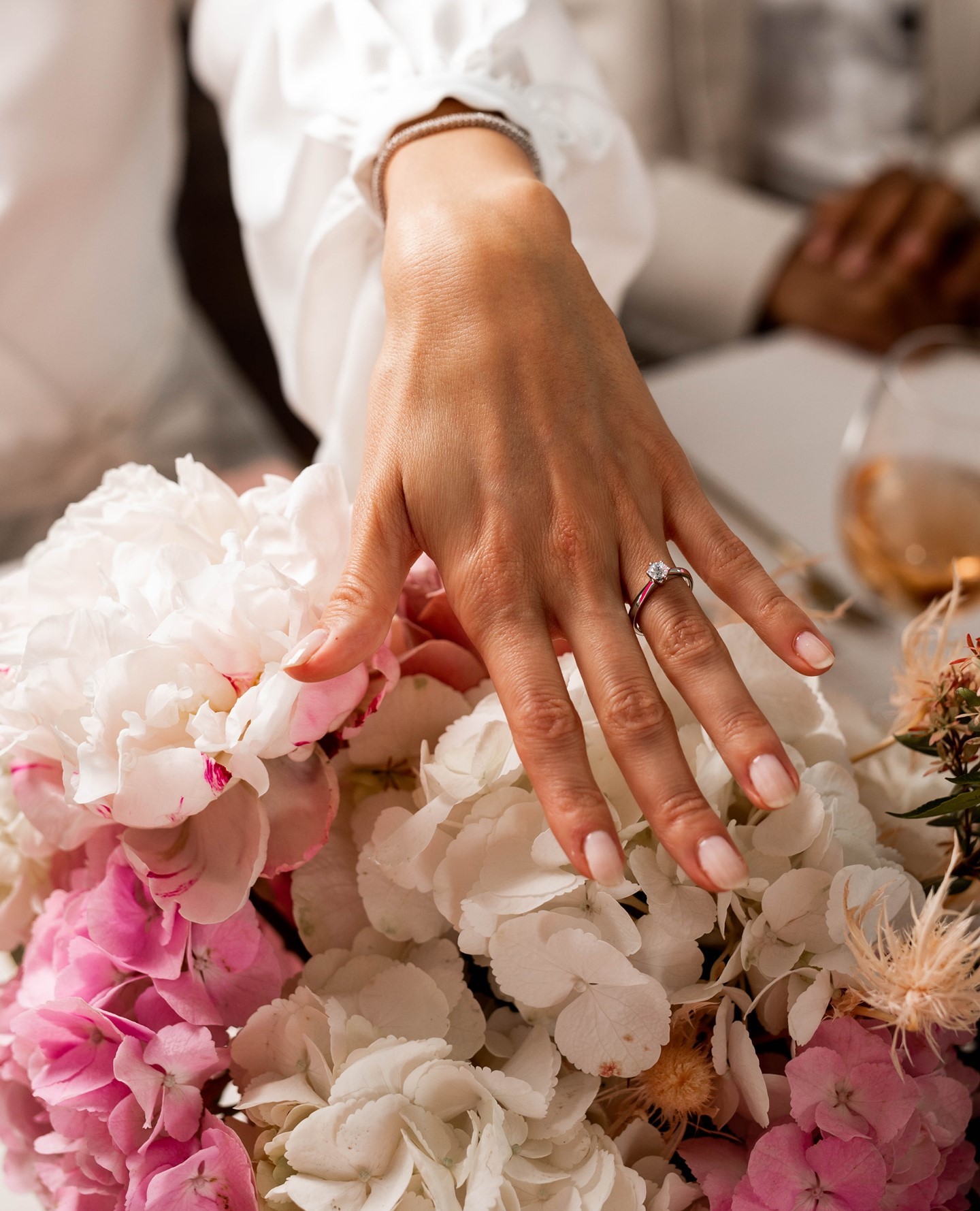 Eine Hand mit einem Ring sitzt über einem bunten Blumenstrauß aus rosa und weißen Blüten, während im Hintergrund verschwommene Personen zu sehen sind.