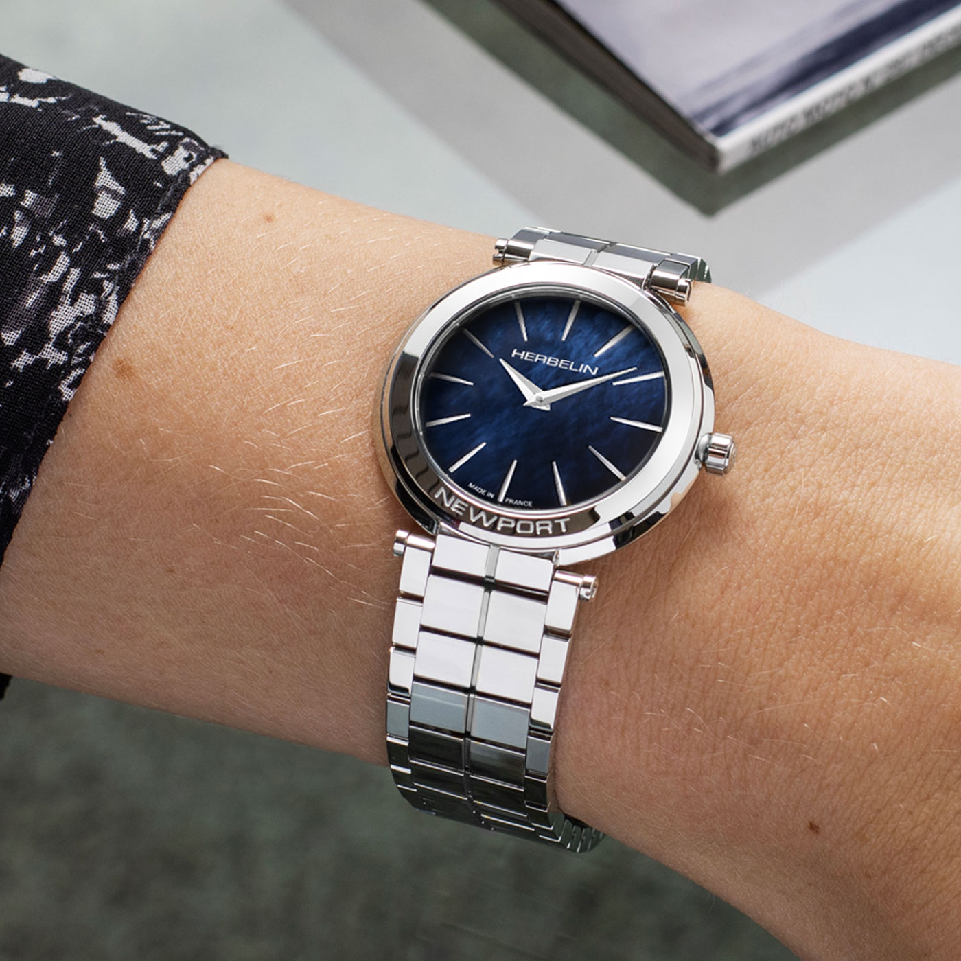 Eine schlanke Armbanduhr mit einem blauen Zifferblatt und einem eleganten Metallarmband liegt an einem Handgelenk, während ein Buch im Hintergrund zu sehen ist.