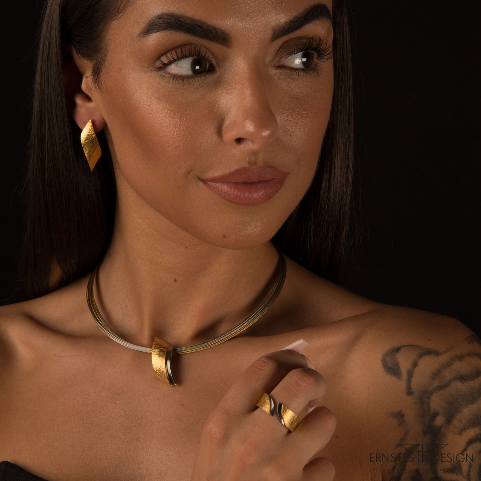 Eine Frau mit langen, dunklen Haaren trägt eleganten Schmuck, darunter Ohrstecker und eine auffällige Halskette. Der Hintergrund ist schwarz.