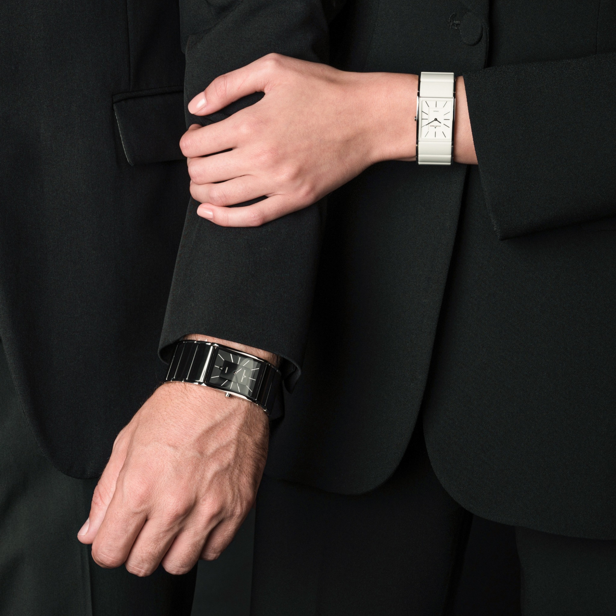 Zwei Personen in formellen Anzügen stehen nebeneinander. Die linke Person trägt eine schwarze Uhr, die rechte eine weiße Uhr, während eine Hand den Arm der anderen umschlingt.
