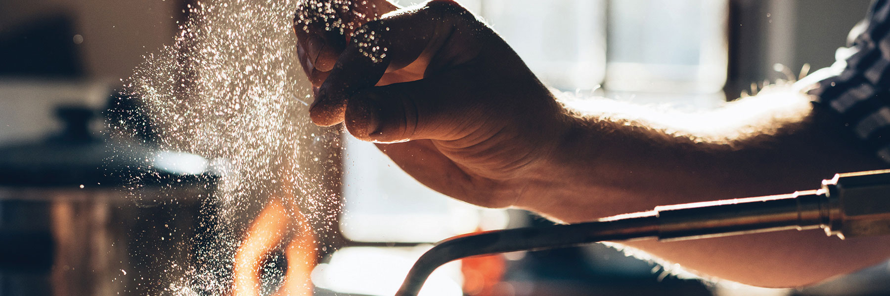 Eine Hand streut feines Pulver über eine offene Flamme, während Sonnenlicht durch ein Fenster strömt und Staubpartikel im Licht sichtbar macht.