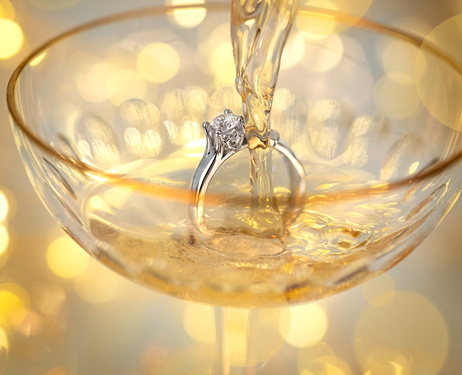 Ein elegantes, funkelndes Champagnerglas mit einem schönen Ring im Inneren, während goldene Flüssigkeit hineinfließt und glitzernde Lichter im Hintergrund leuchten.