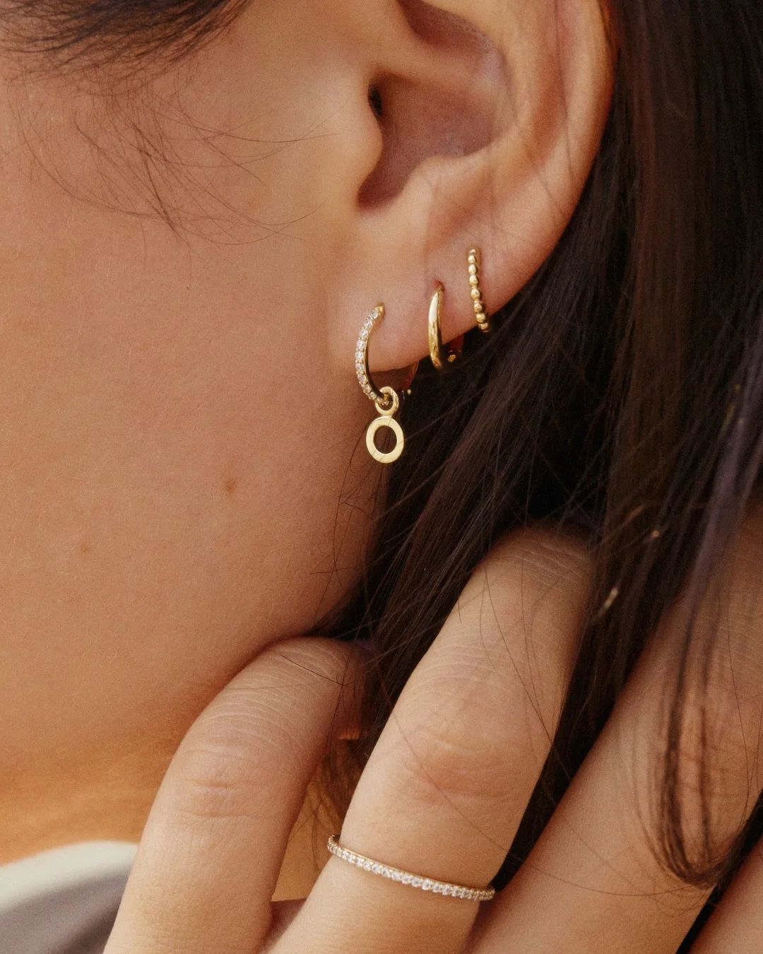 Nahaufnahme eines Ohres mit mehreren goldenen Ohrringen, darunter ein kleiner mit einem Anhänger in Form eines Kreises, und einer zarten Ringed.