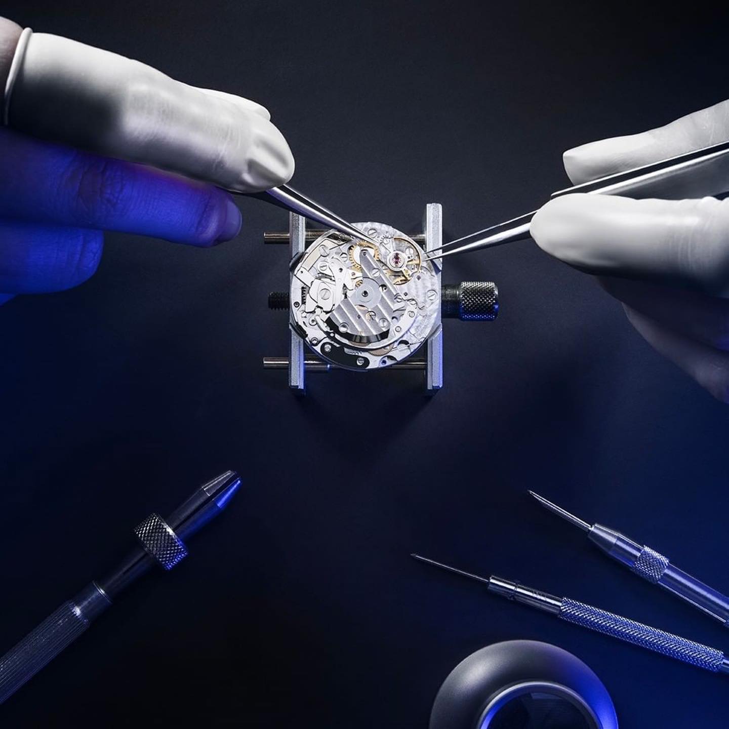 Eine Person mit weißen Handschuhen arbeitet präzise an einem Uhrenmechanismus, umgeben von Werkzeugen wie Pinzetten und einem Maßstab auf einem dunklen Hintergrund.
