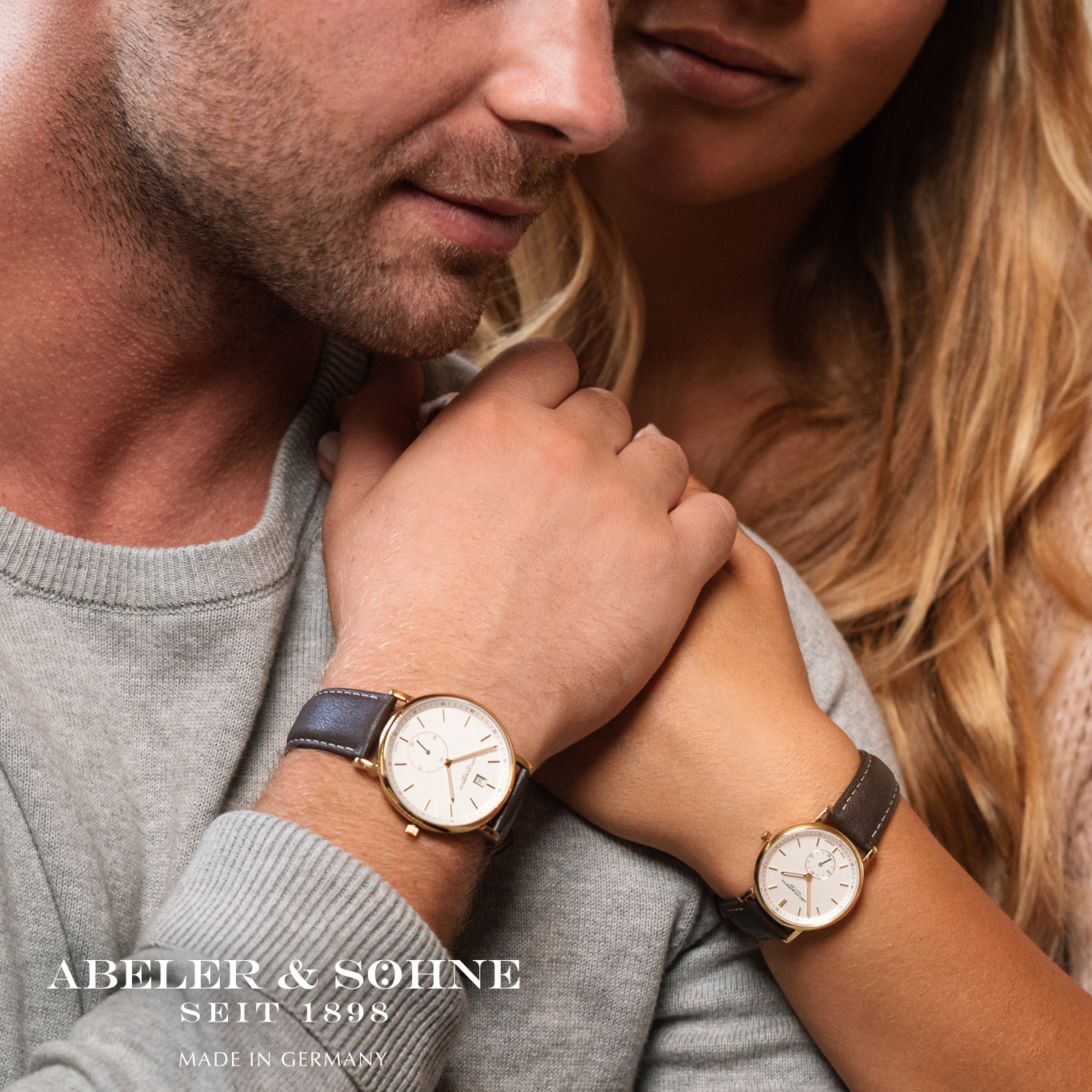 Ein Mann und eine Frau zeigen ihre Uhren. Der Mann trägt eine goldene Uhr mit grauem Lederarmband, die Frau ebenfalls eine goldene Uhr mit schwarzem Armband. Beide lächeln und stehen nah beieinander.