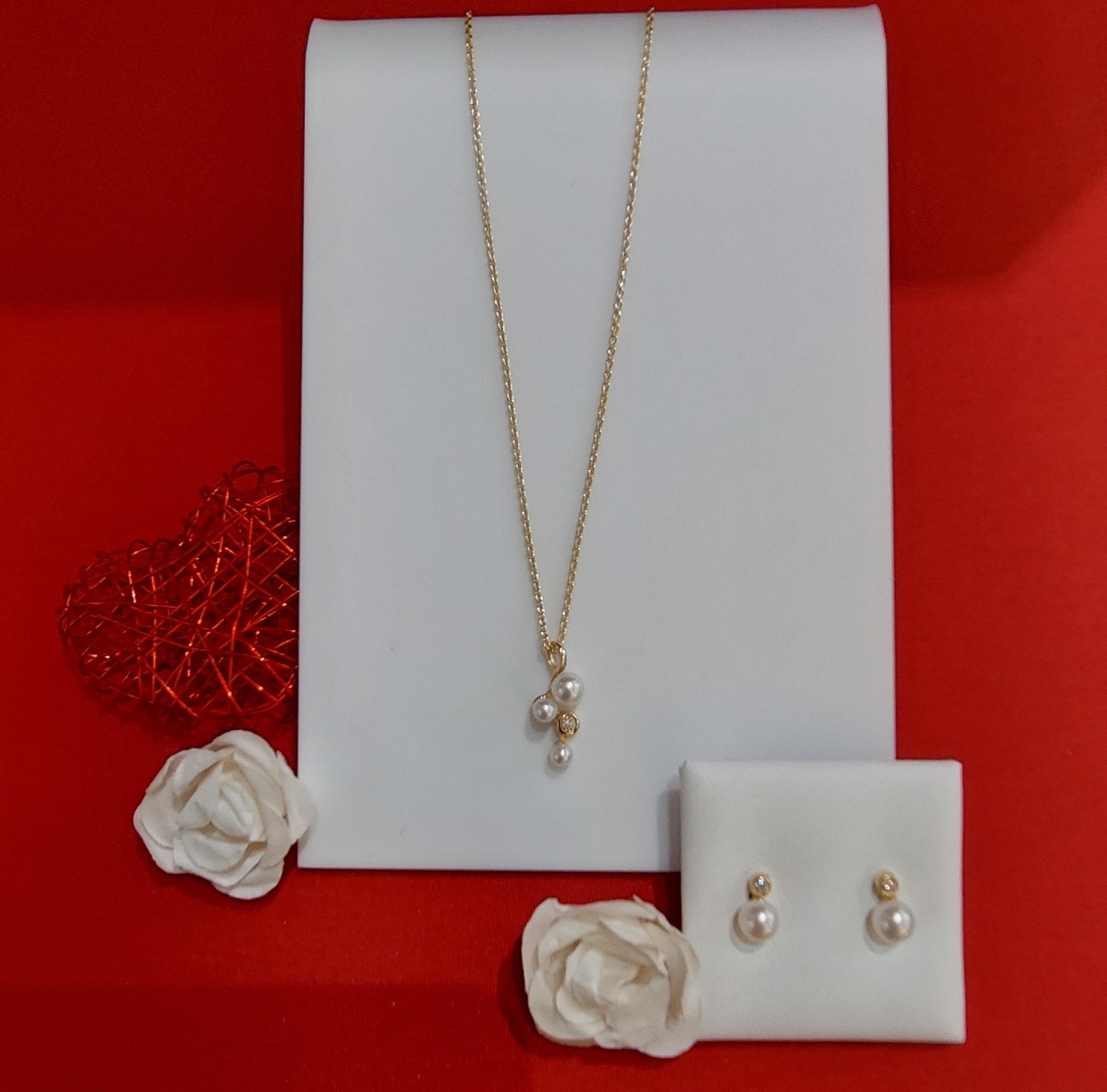 Eine goldene Kette mit einer Perlenskulptur hängt an einem weißen Display, neben einem Paar Perlenohrringe und einigen Papierrosen auf rotem Hintergrund.