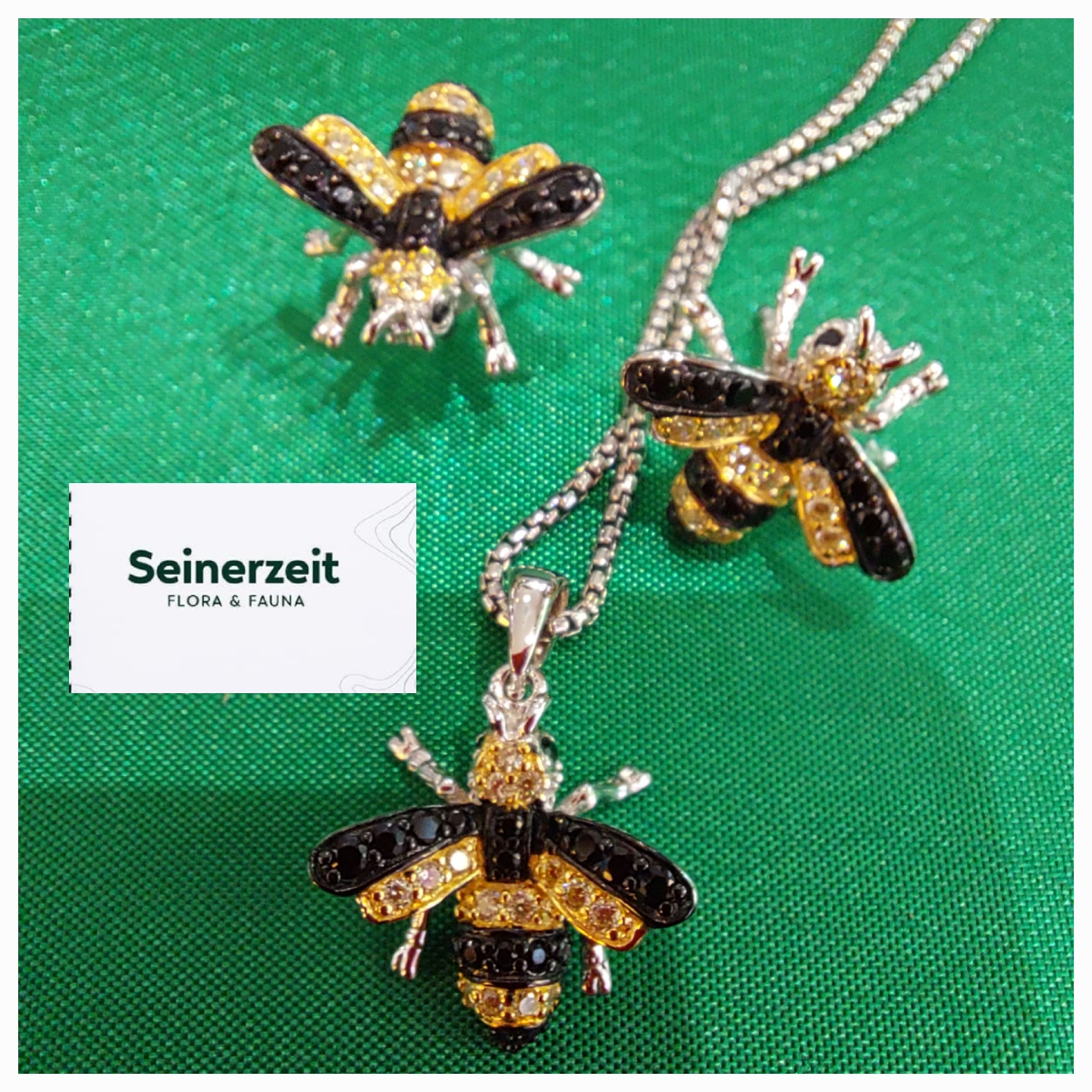 Drei schmucke Bienenanhänger aus glänzendem Material liegen auf einem grünen Untergrund. Ein Logo von 