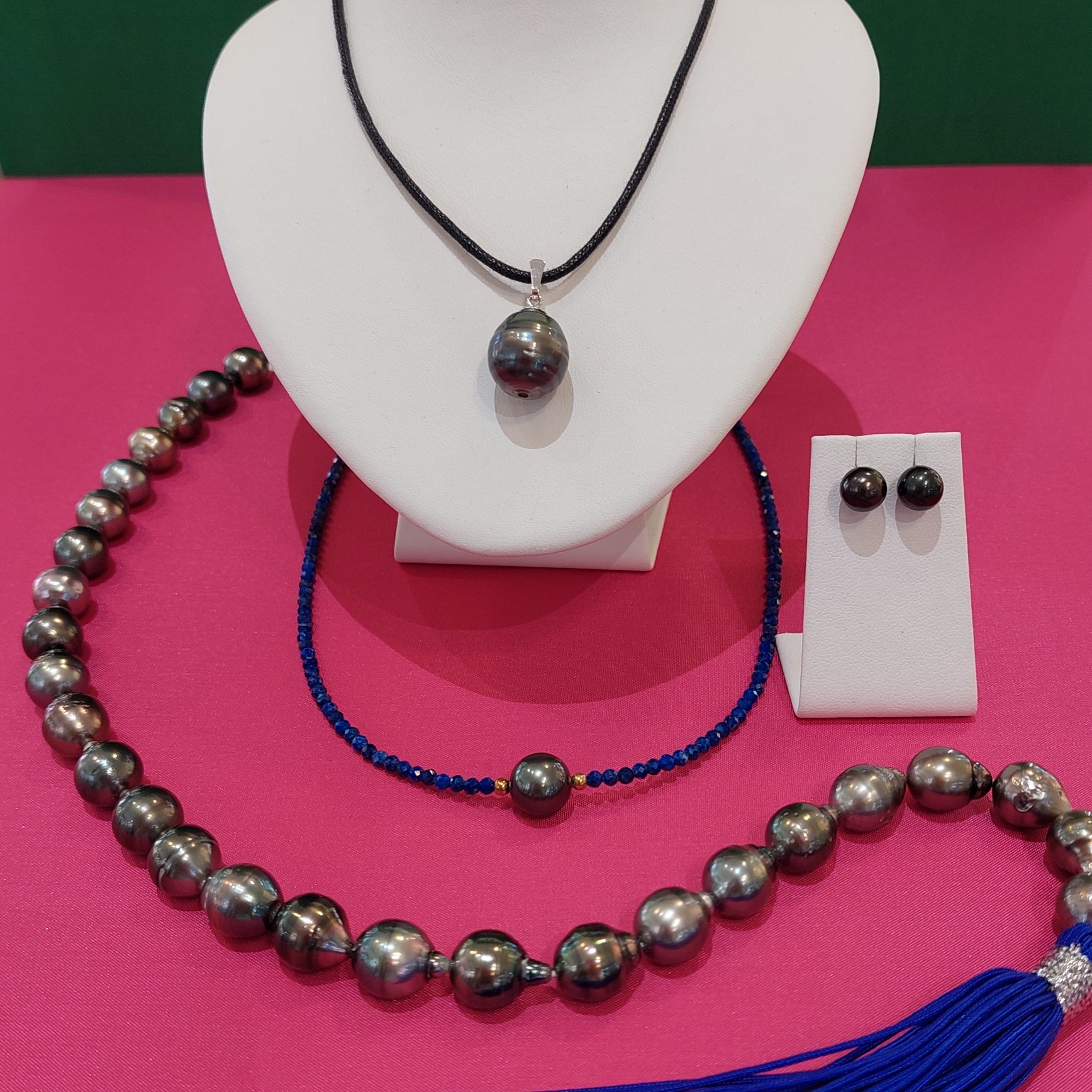 Ein Schmuckdisplay mit einer Halskette aus großen Perlen, einem blauen Armband, Ohrringen und einer Kette mit einem Anhänger. Der Hintergrund ist pink.