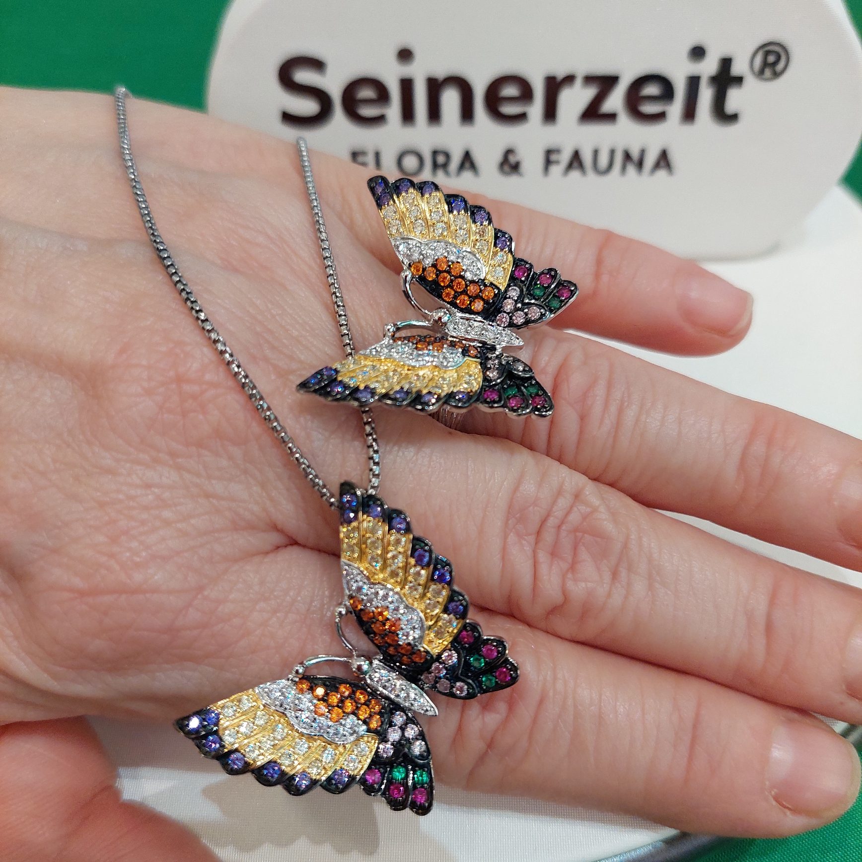 Eine Hand mit zwei kunstvoll gestalteten Schmetterlingen als Schmuckstücken, umgeben von einem Hintergrund mit der Aufschrift 