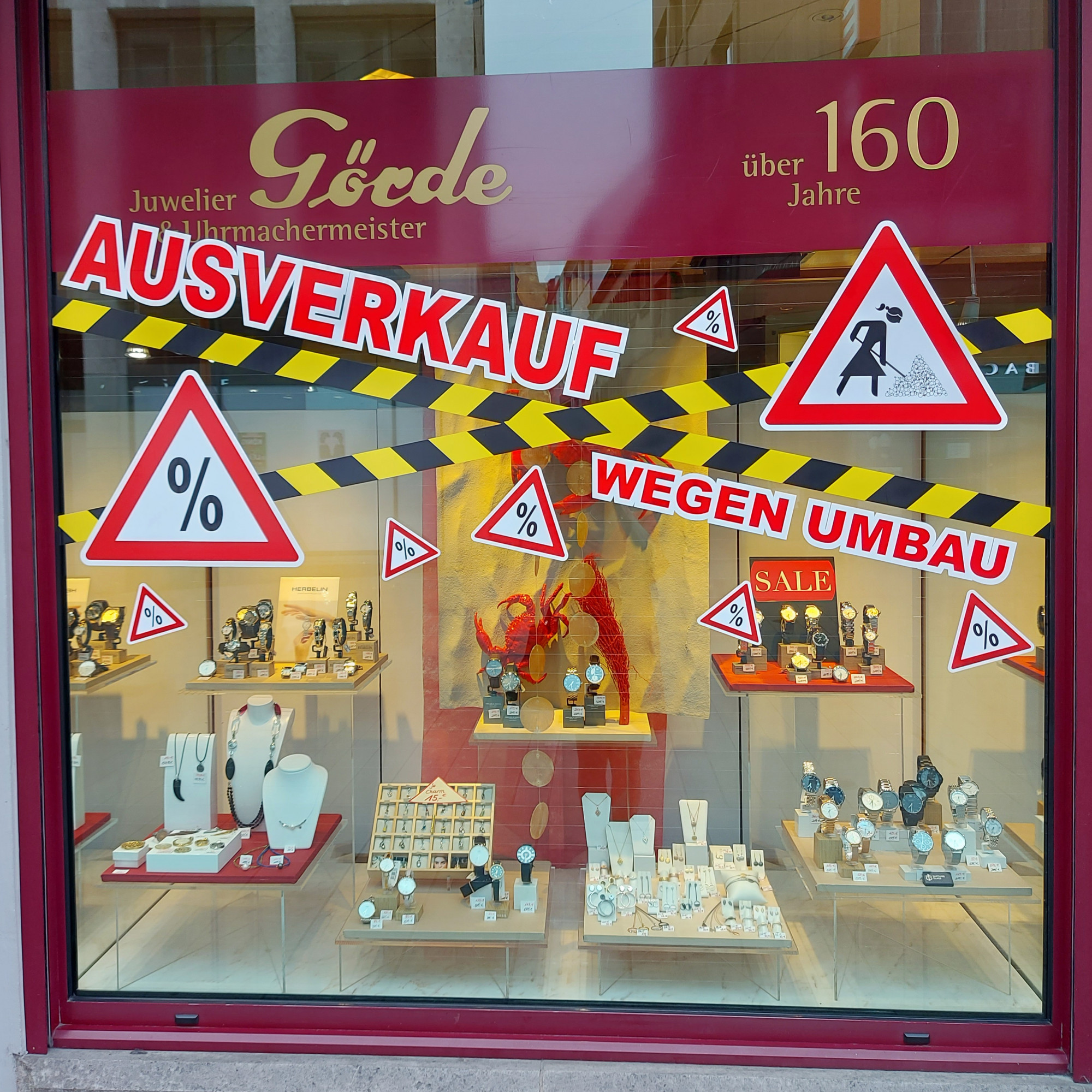 Im Schaufenster eines Juweliers sind Uhren und Schmuck ausgestellt. Bunte Schilder verkünden „Ausverkauf wegen Umbau“ und weisen auf Rabatte hin.