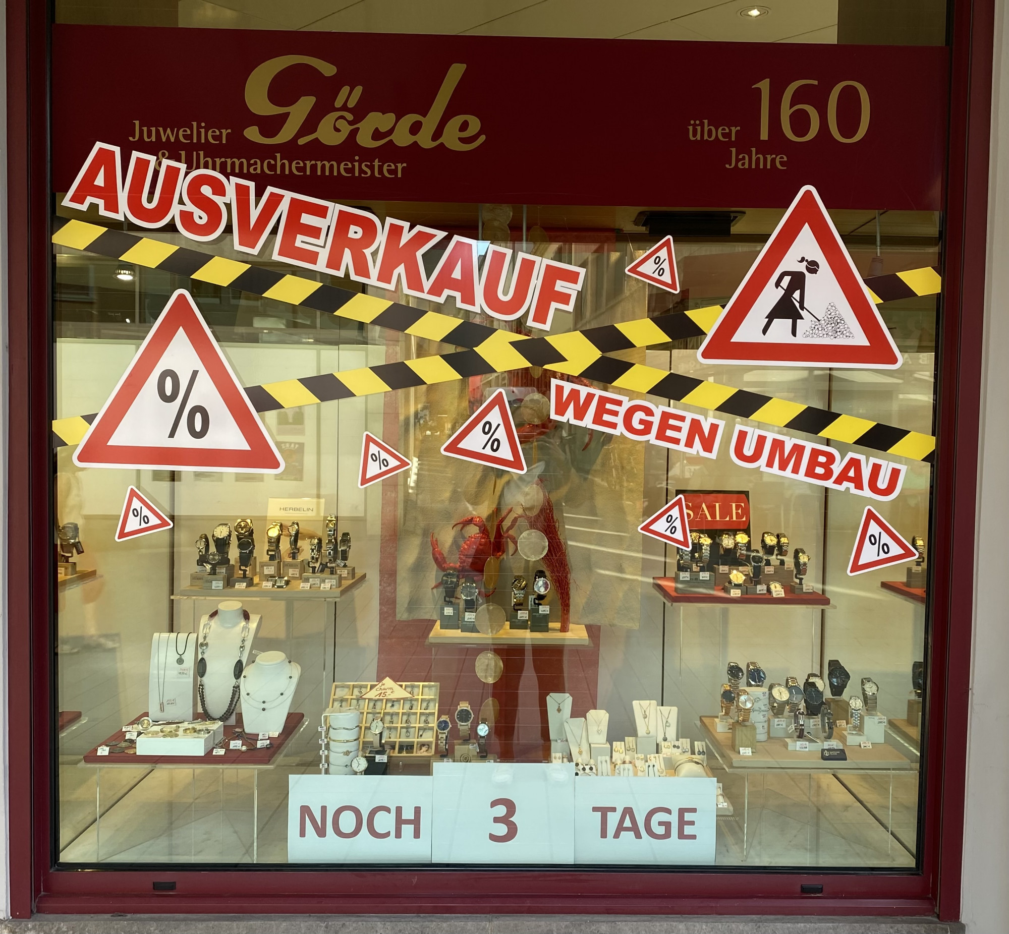 Der Schaufensterbereich eines Juweliers zeigt Uhren und Schmuckstücke, umgeben von roten Warnzeichen. Es gibt große Schriftzüge „AUSVERKAUF“ und „WEGEN UMBLAU“, sowie „NOCH 3 TAGE“.