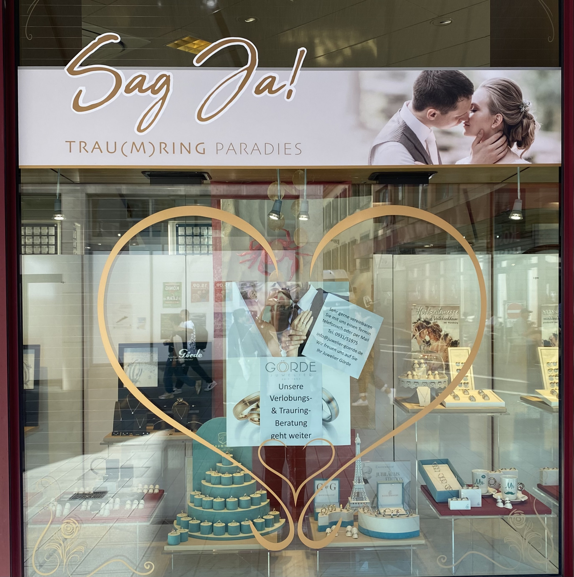 In einem Schaufenster wird die Trauring-Ausstellung präsentiert. Ein Herzrahmen umrahmt den Inhalt, mit Ringen und Schmuckstücken sowie einer Beratungshinweis.