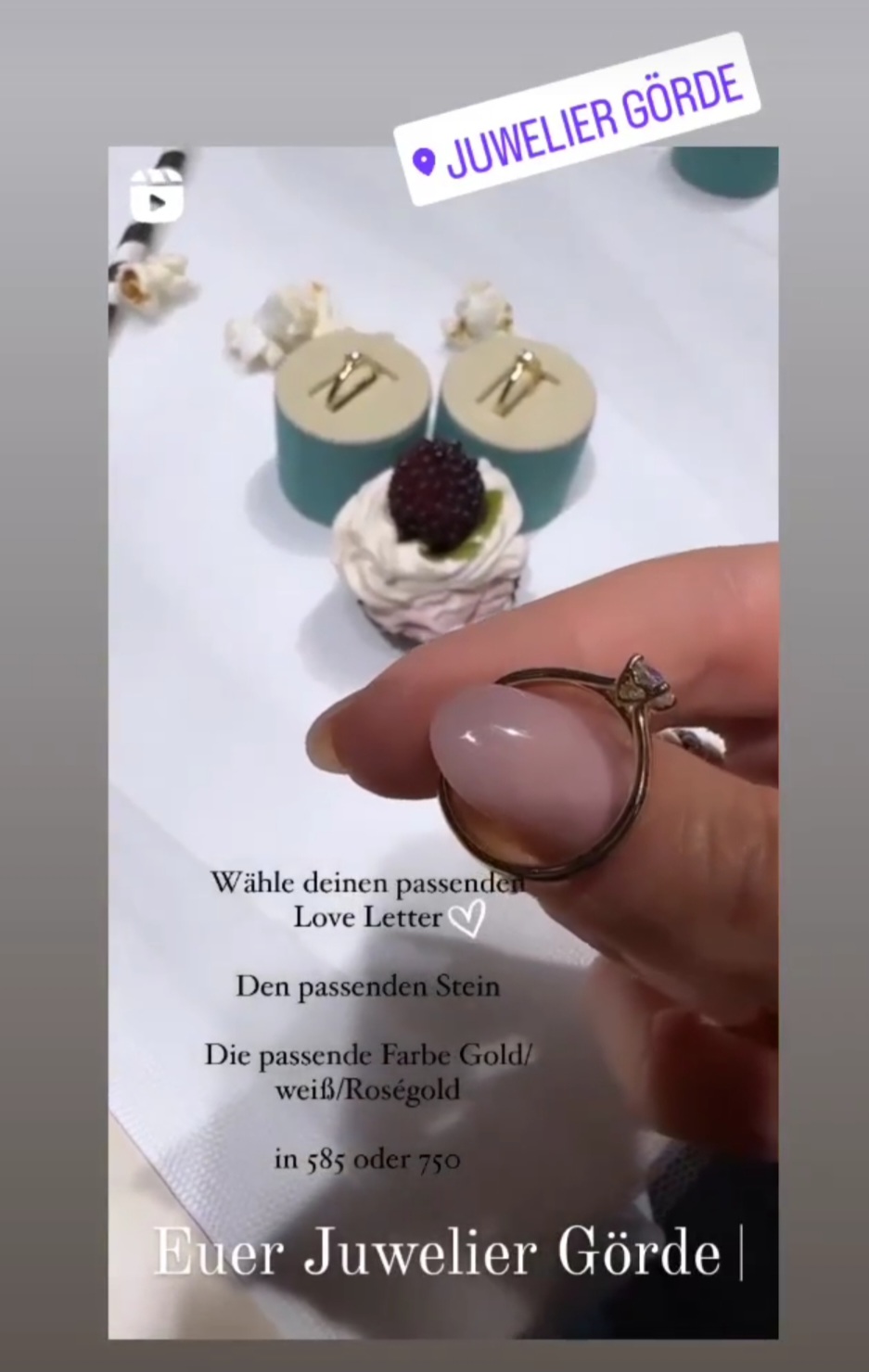 Eine Hand hält einen eleganten Ring mit einem Stein. Im Hintergrund sind zwei Schmuckkästchen und ein Cupcake mit Beeren zu sehen. Textinformationen erscheinen.