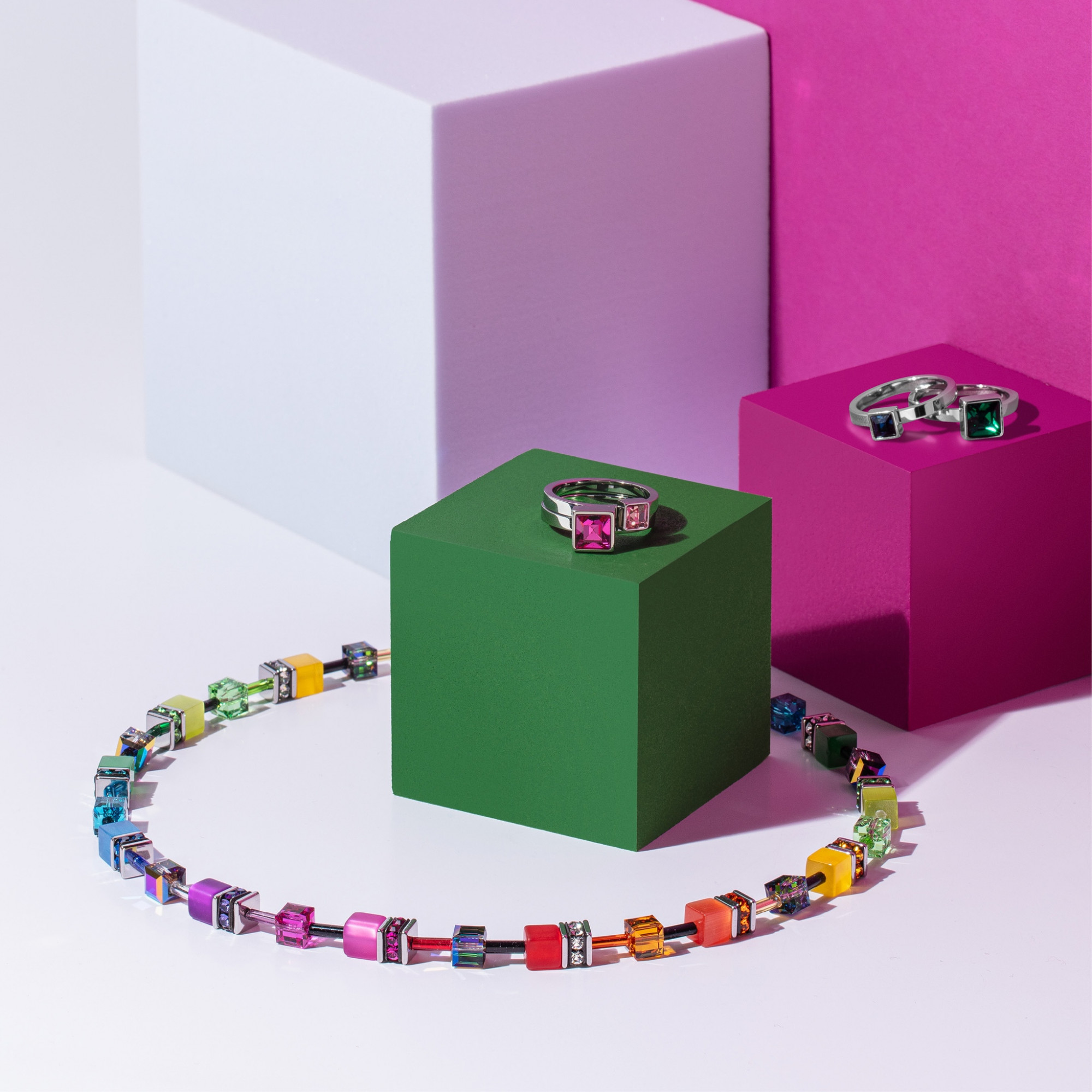 Ein mehrfarbiger Schmuck besteht aus einer Kette und Ringen, die auf grünen, rosa und weißen Würfeln präsentiert werden. Bunte Steine ziehen die Aufmerksamkeit an.